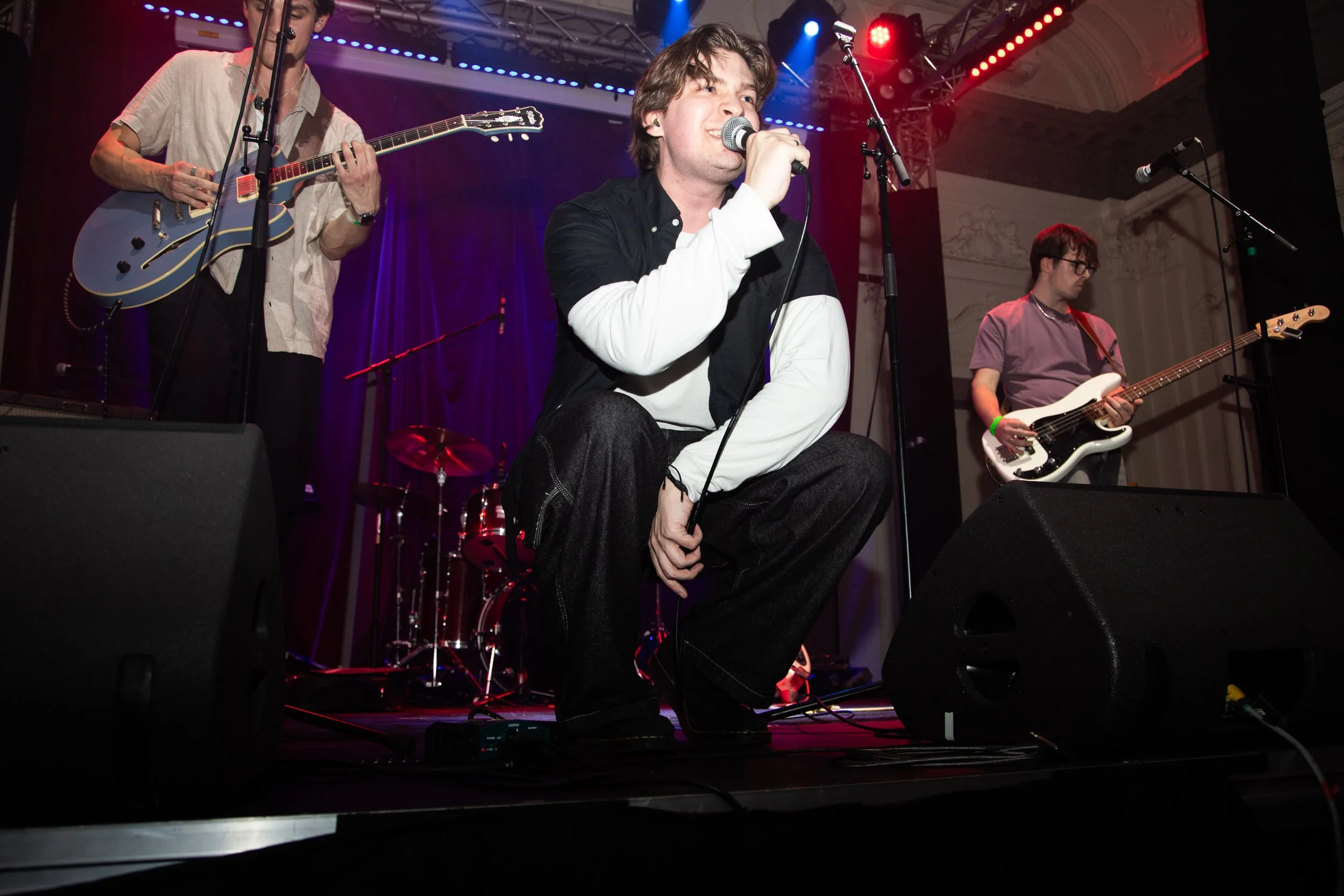 A band performing on stage with a male singer kneeling and holding a microphone, a guitarist on the left, and a bassist on the right, with colorful stage lights and microphones around them.