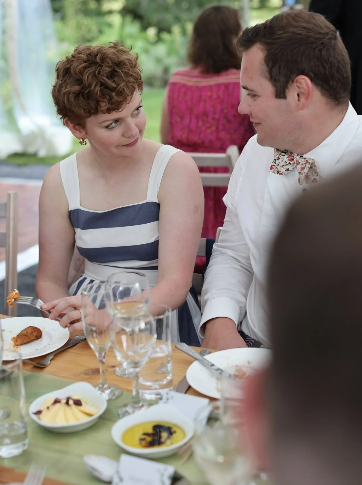 A woman with short curly red hair and a striped sleeveless dress looks at a man with short dark hair and a floral bow tie as they sit at a table during a meal. The table has plates, glasses, and bowls with food and drinks, and is set in an outdoor se