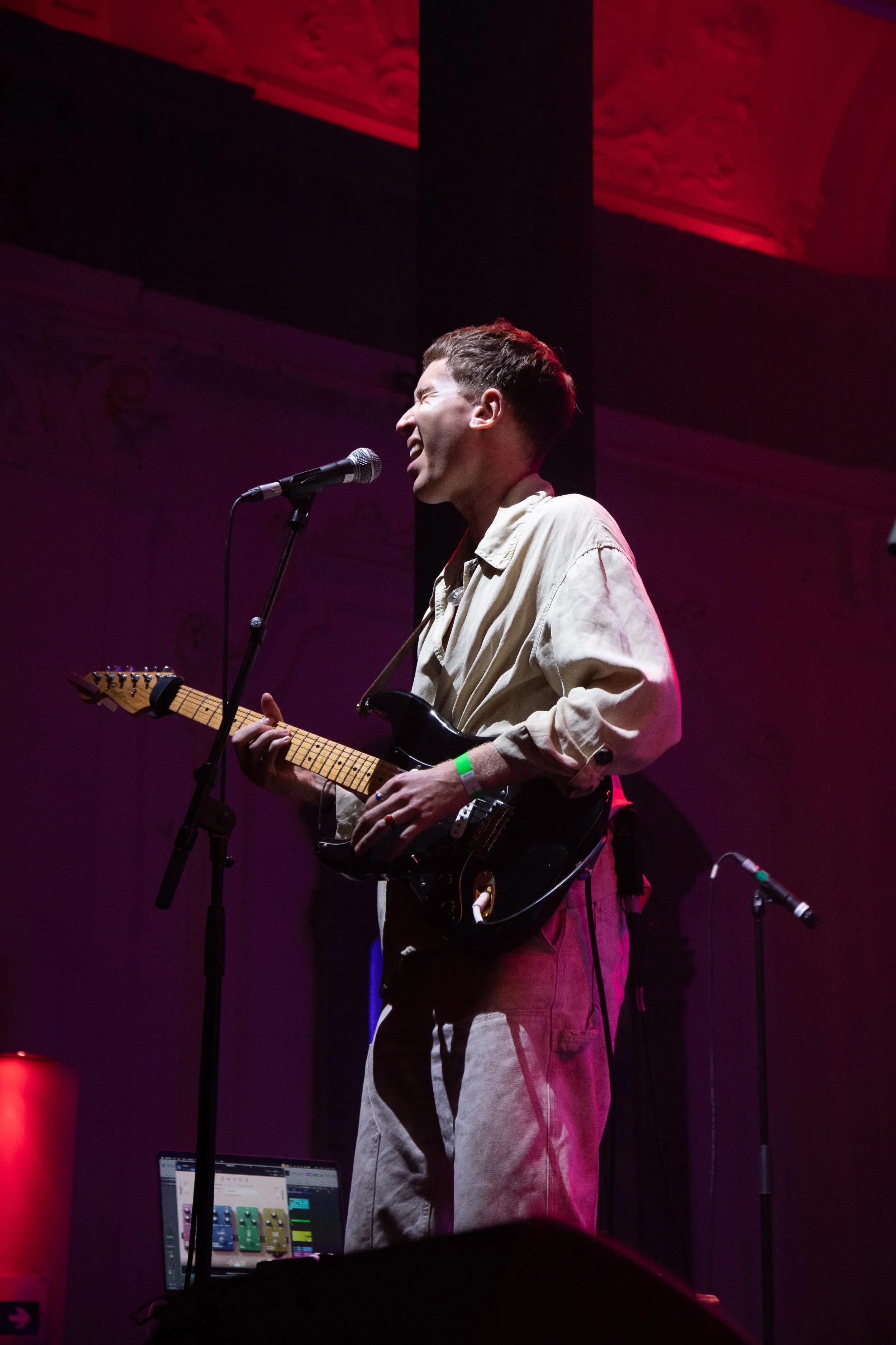 A male musician with short hair is singing into a microphone while playing an electric guitar on stage, with a computer and music equipment visible in the background, under colorful stage lighting.