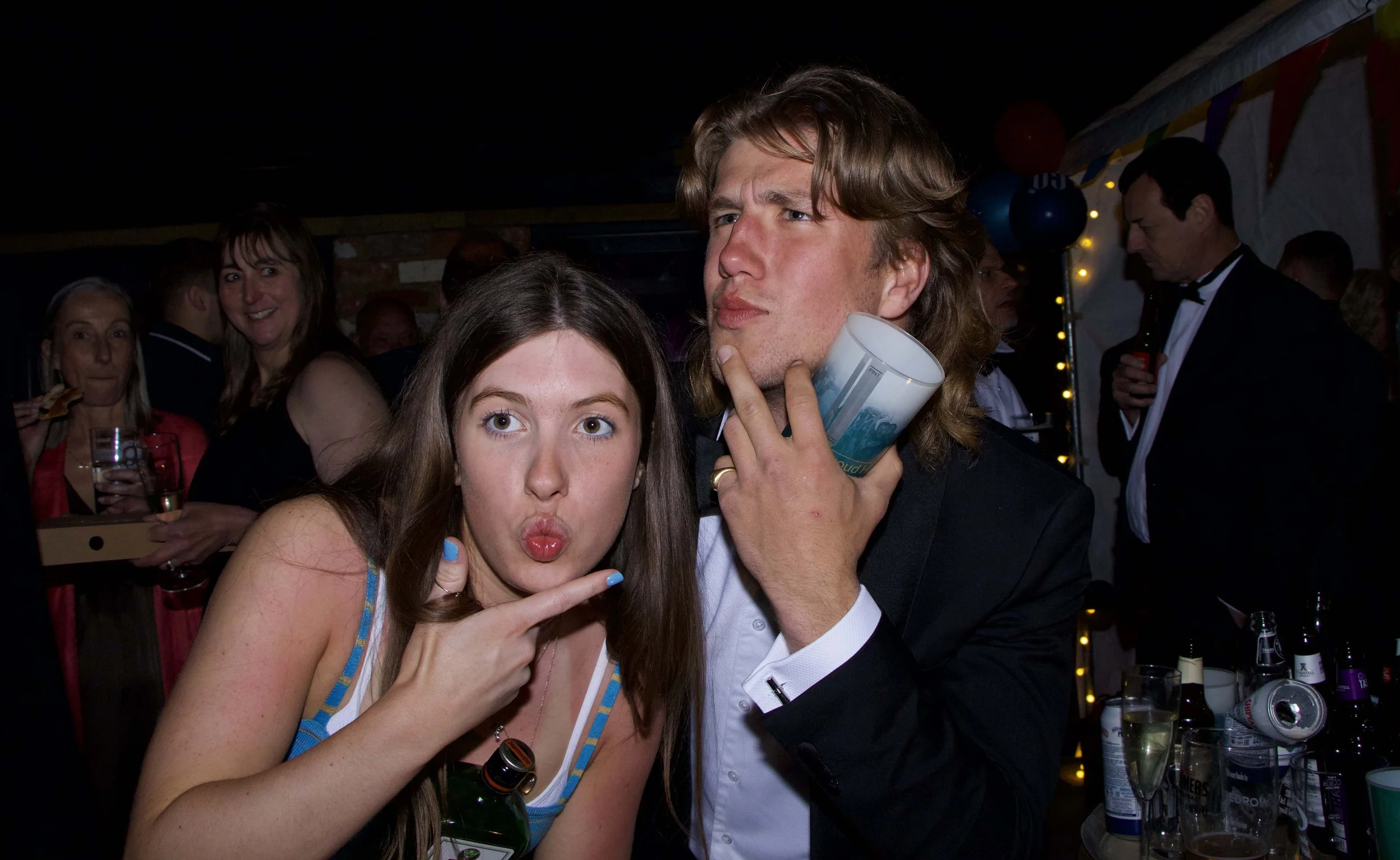 A young woman with long brown hair making a duck face and a young man with long hair and a suit making a thoughtful face, both posing at a party with other people in the background.