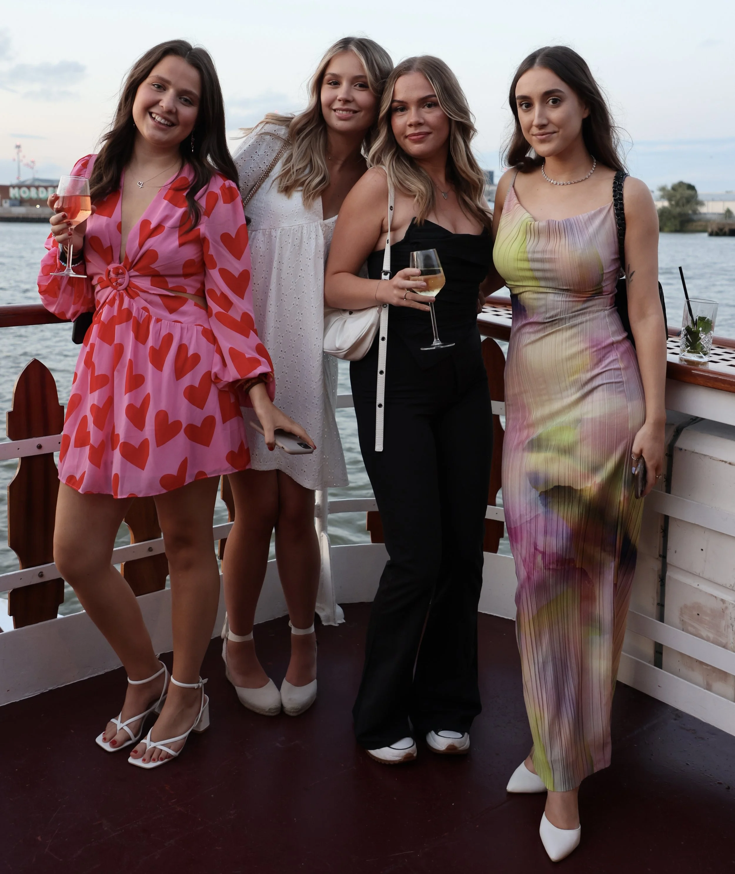 Four women standing on a boat deck near water, smiling at the camera. They are dressed in fashionable outfits, holding drinks, with a background of water and sky.