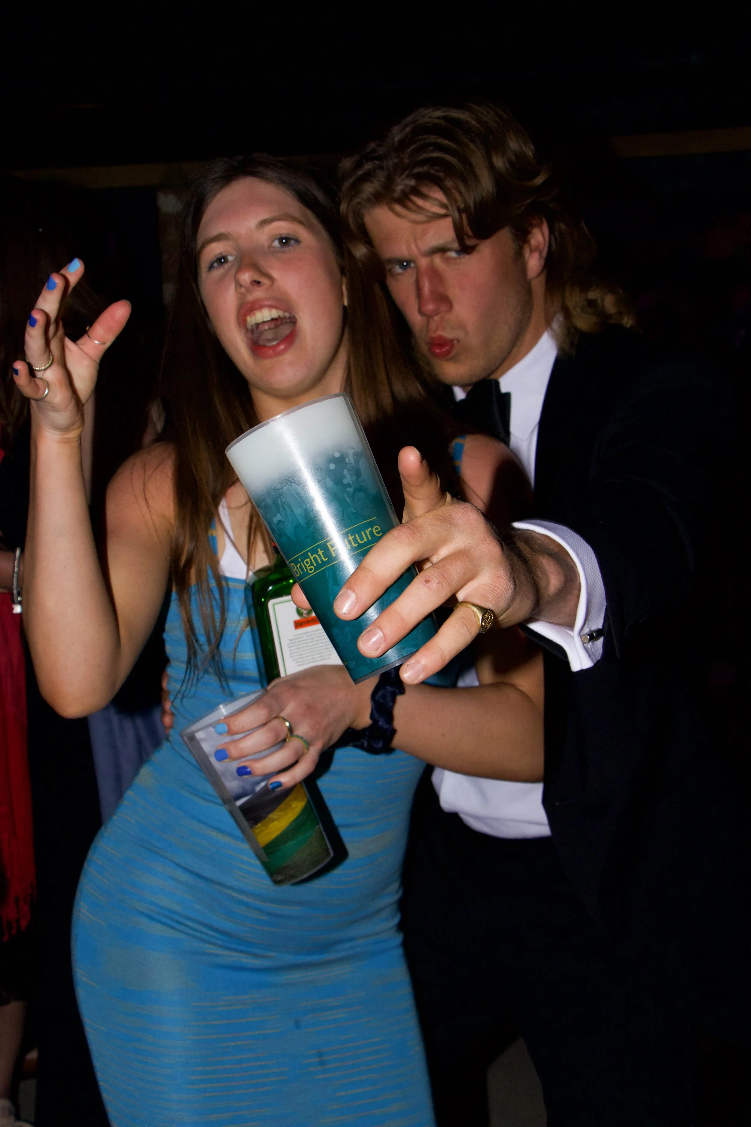Two people, a woman and a man, posing together at a social event, holding a drink, with the woman wearing a blue striped dress and the man in a tuxedo, both making expressive gestures.