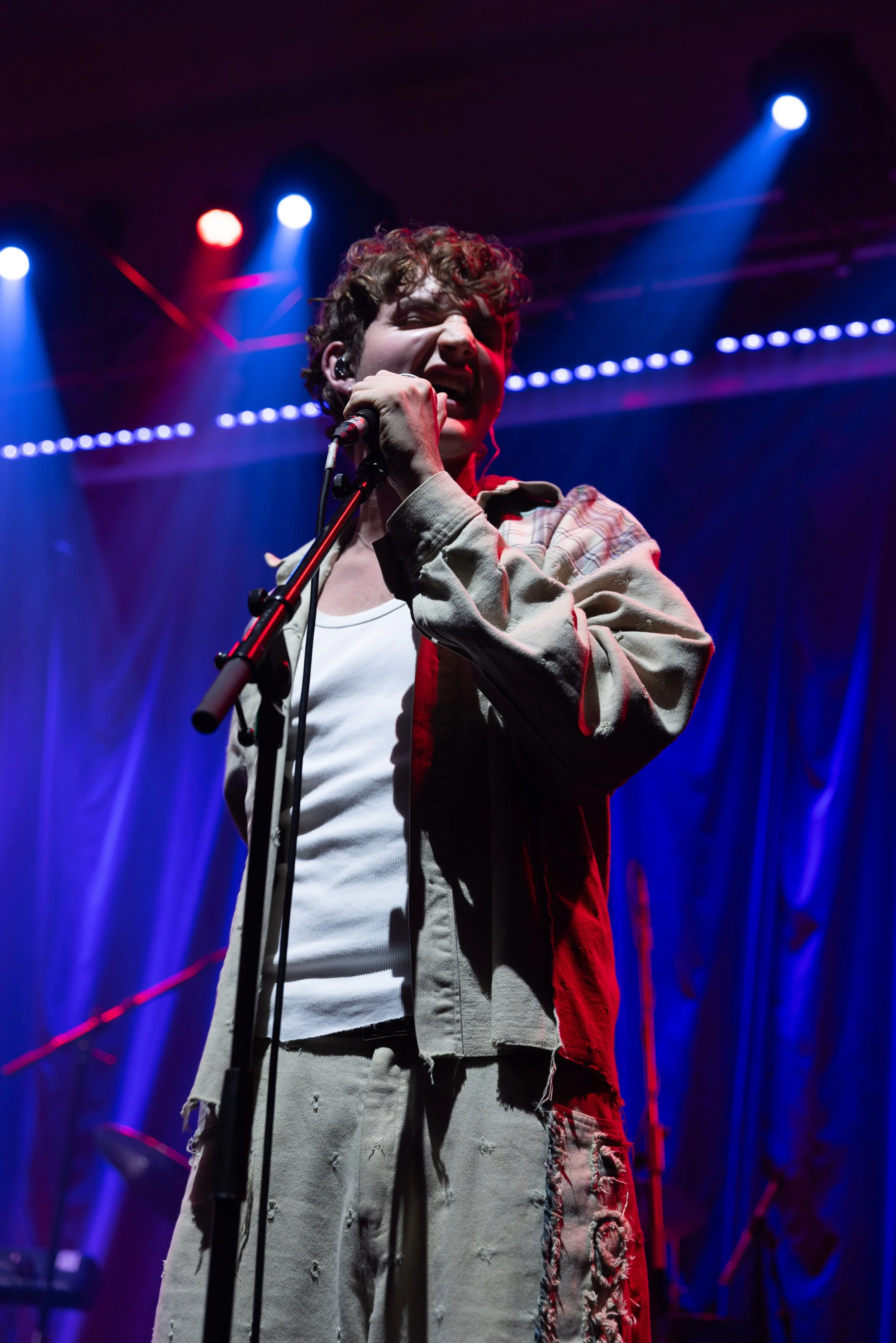 A young man with curly hair singing into a microphone on stage, illuminated by blue and red stage lights, with a dark background.