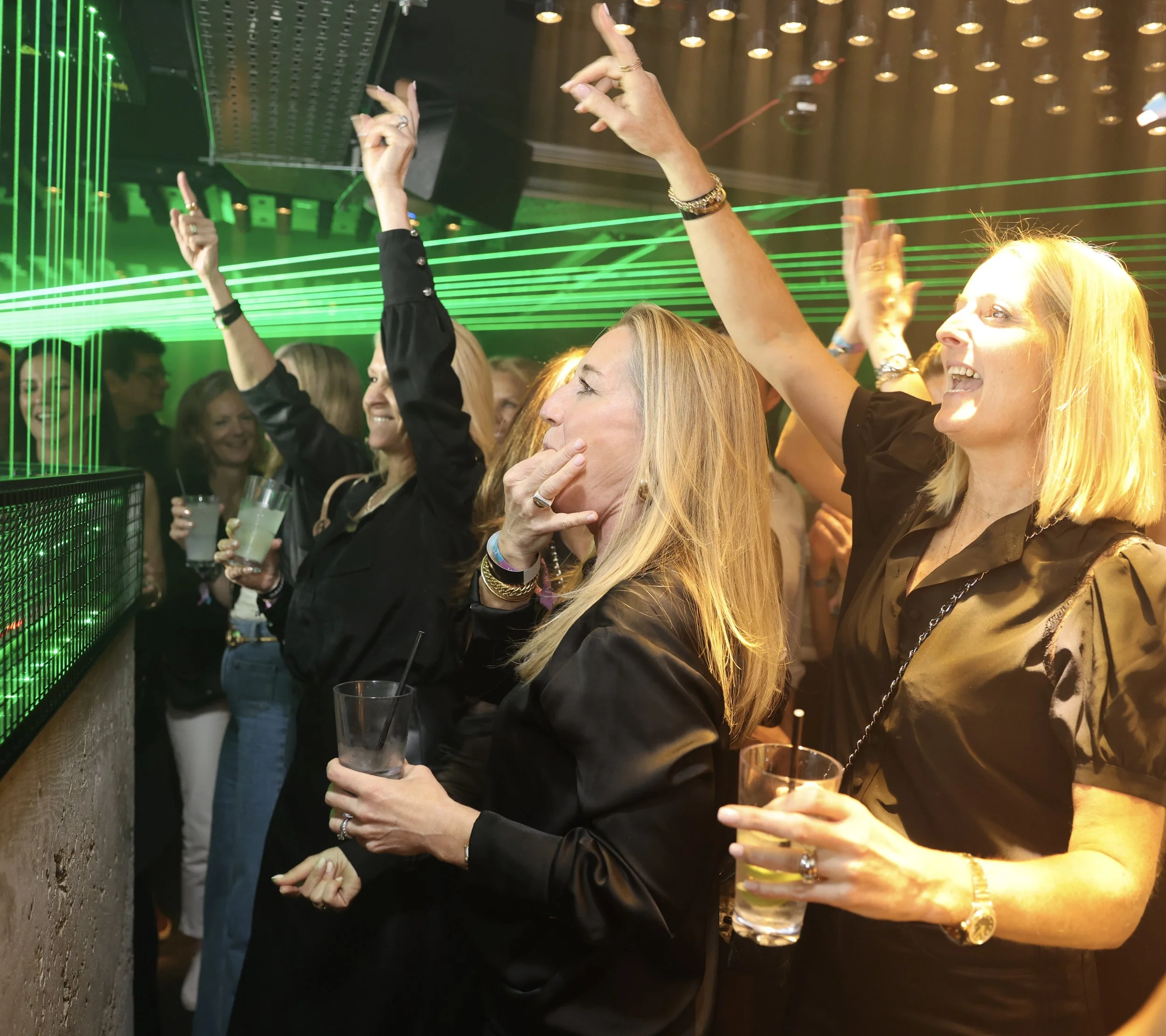 People dancing and socializing in a nightclub with green laser lights and warm yellow lighting, holding drinks, enjoying the music and lively atmosphere.