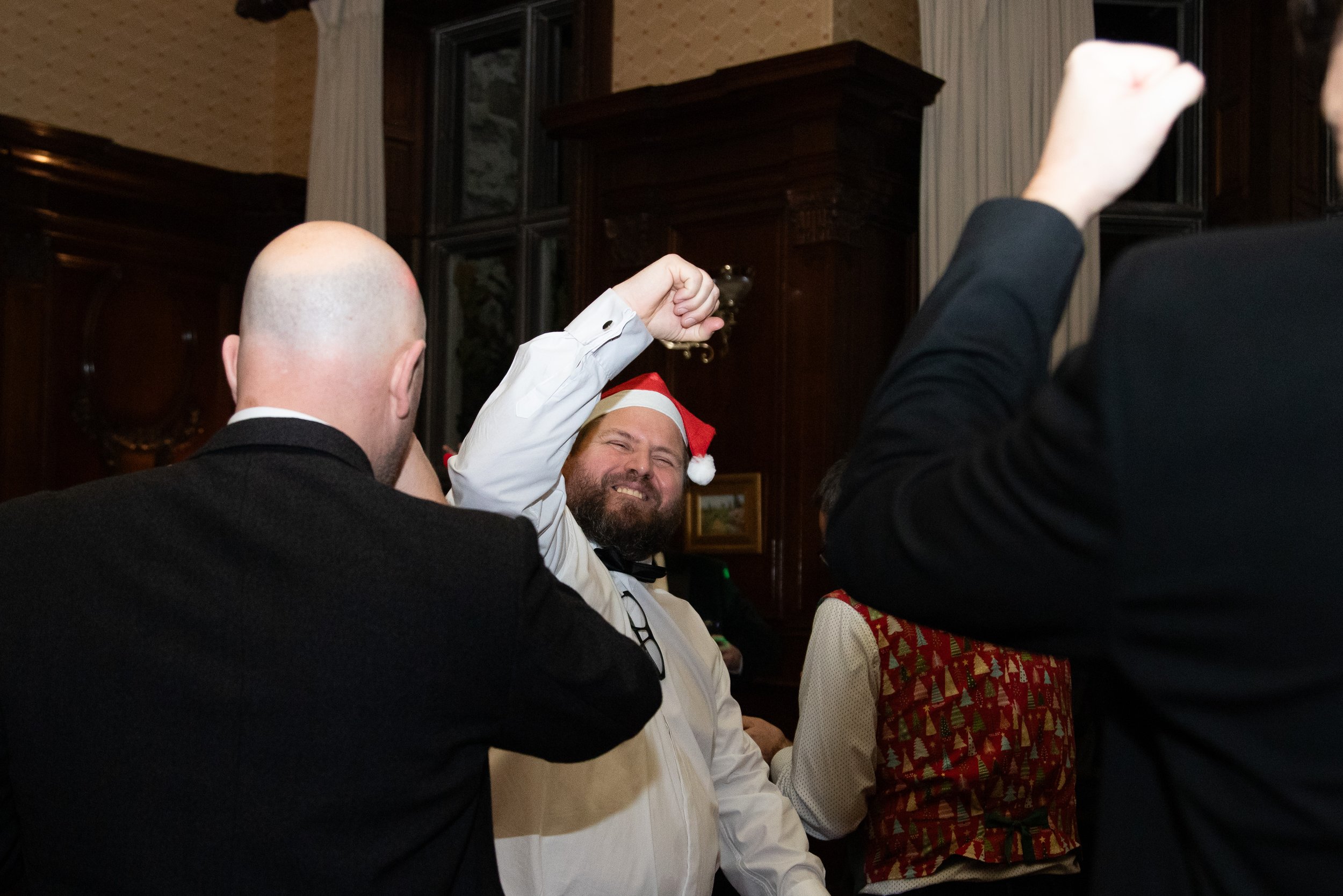 A man wearing a Santa hat and a tuxedo is smiling and raising his hand in a fist bump during a Christmas celebration, with others around him in formal attire.
