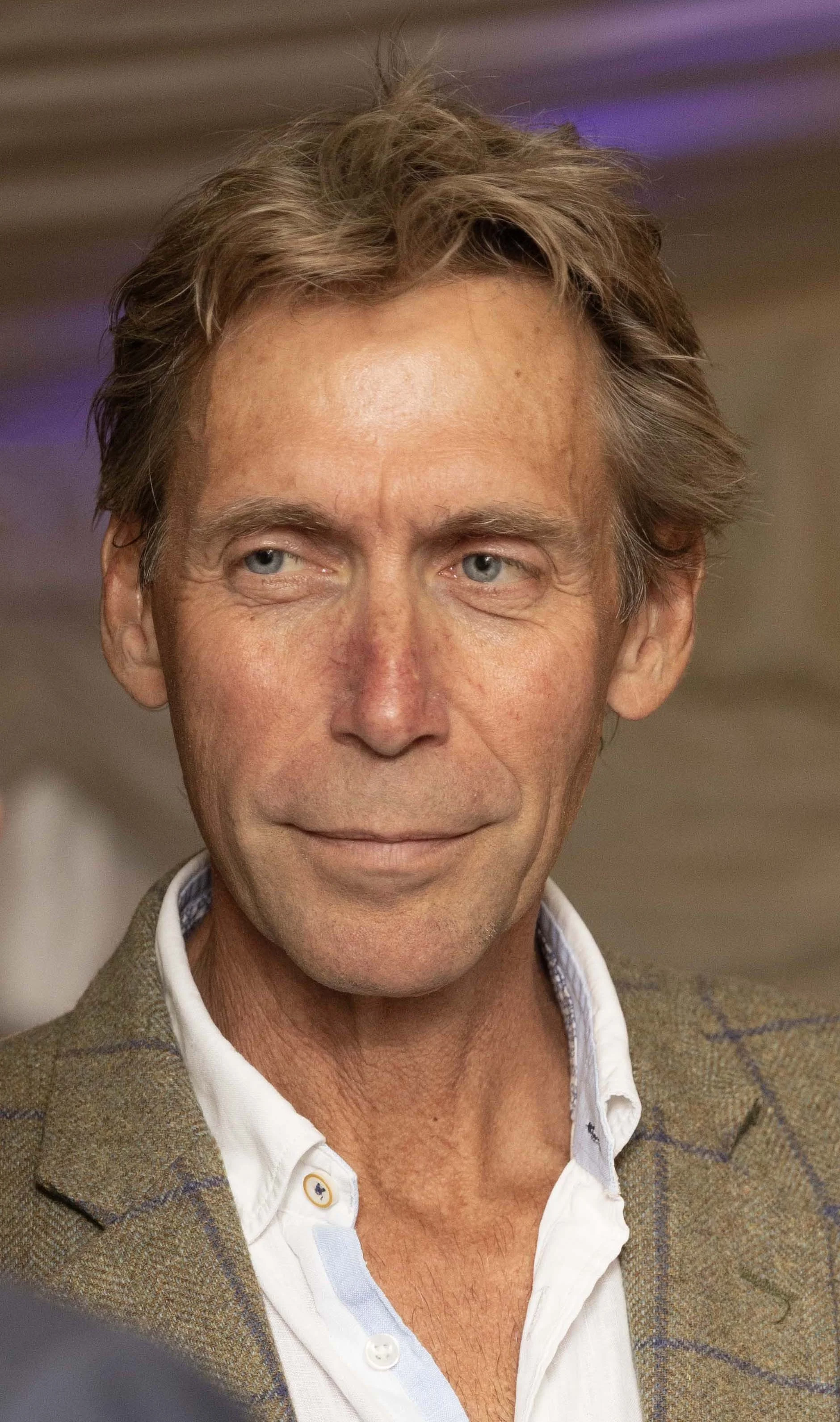 Close-up of a middle-aged man with light brown hair, blue eyes, and fair skin, wearing a brown plaid blazer over a white shirt.