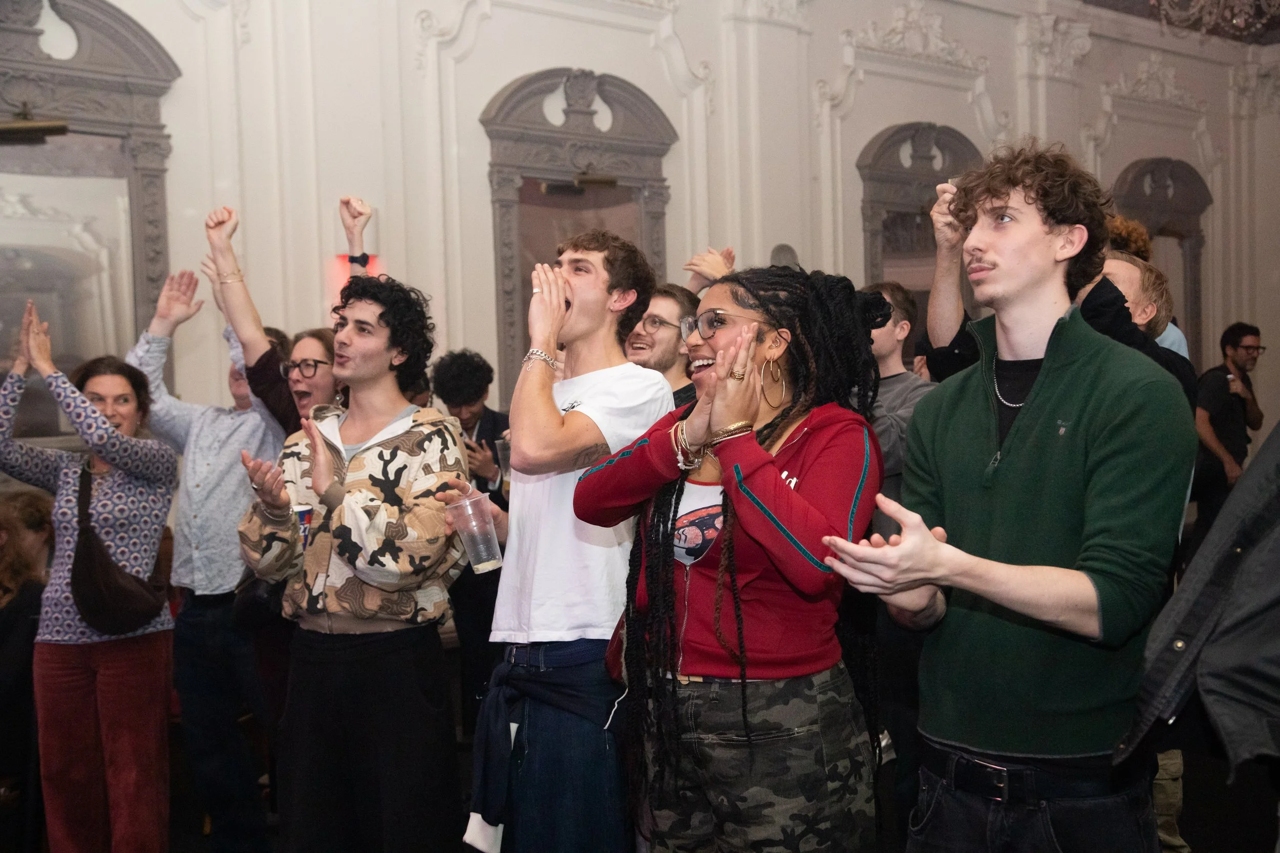 A crowd of people standing and watching at an indoor event, some smiling and cheering.