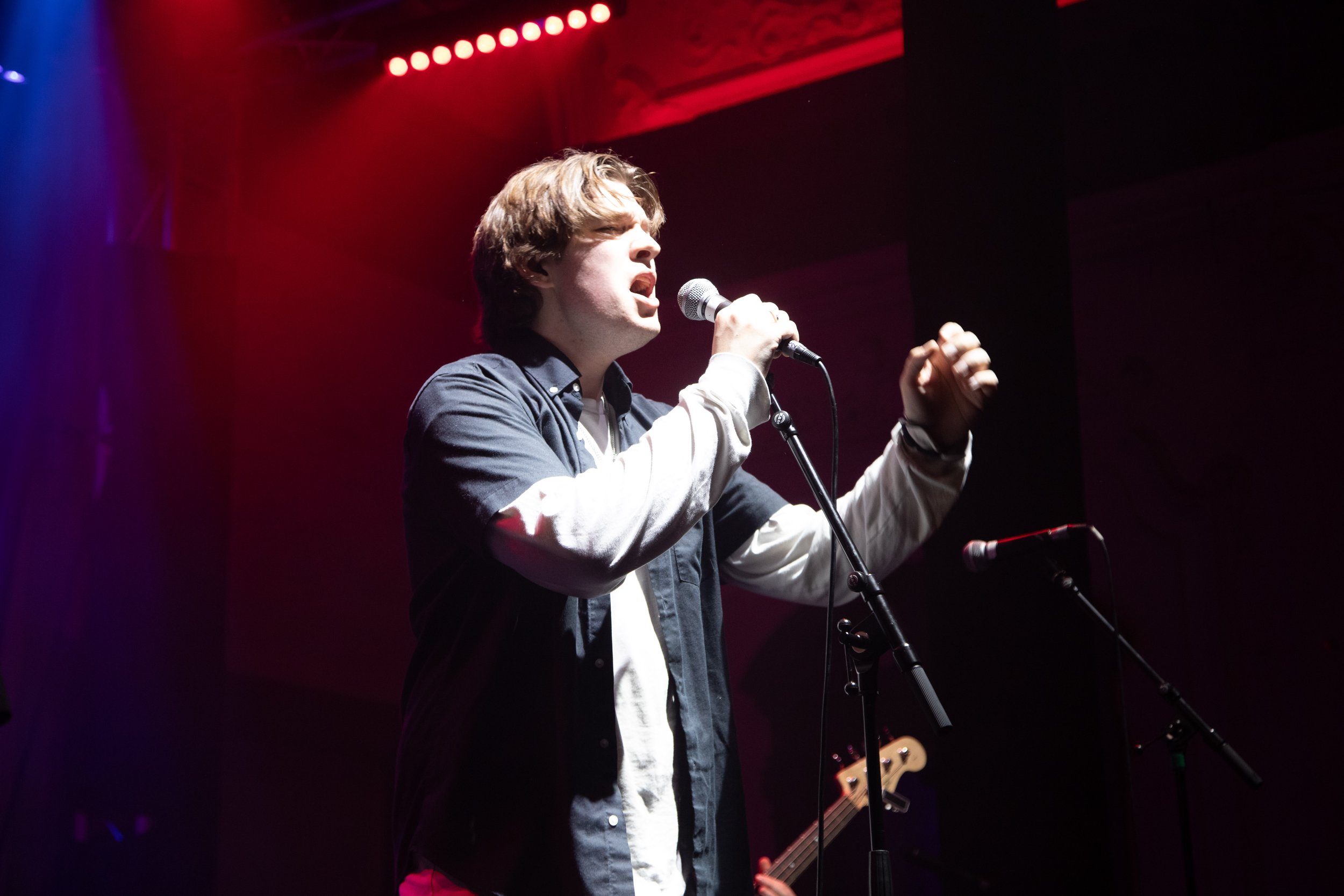 A young man singing into a microphone on stage with stage lights overhead, wearing a dark jacket and a white shirt with rolled sleeves.