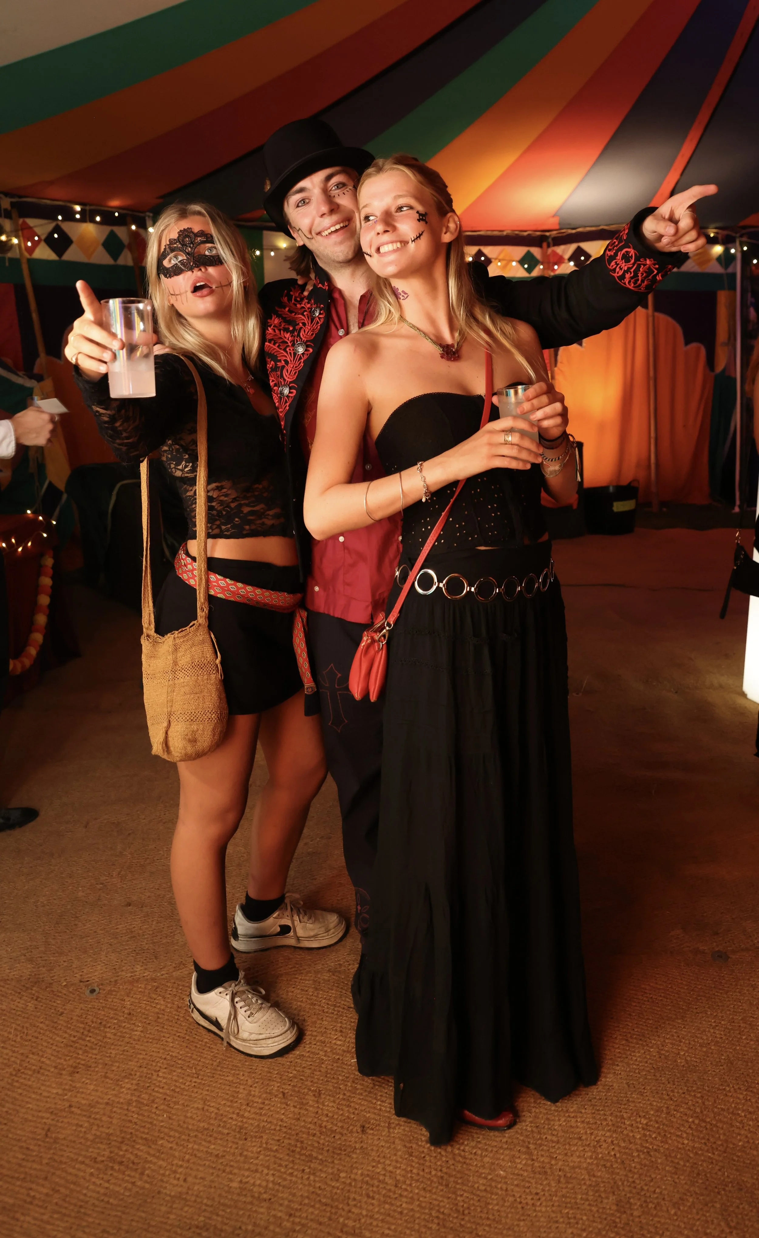 Three people at a festive event, dressed in costumes with face paint and accessories, smiling and posing together under a colorful tent with string lights in the background.