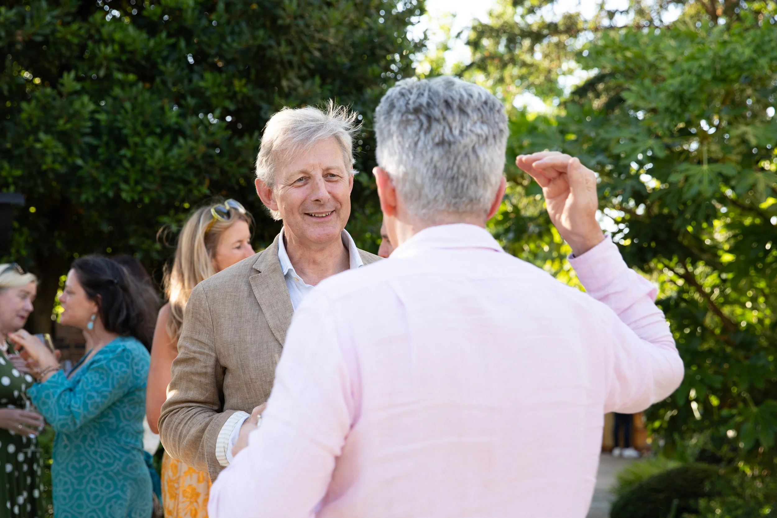 Two older men with gray hair are talking and greeting each other outdoors at a gathering, with a group of people chatting and standing among green trees in the background.