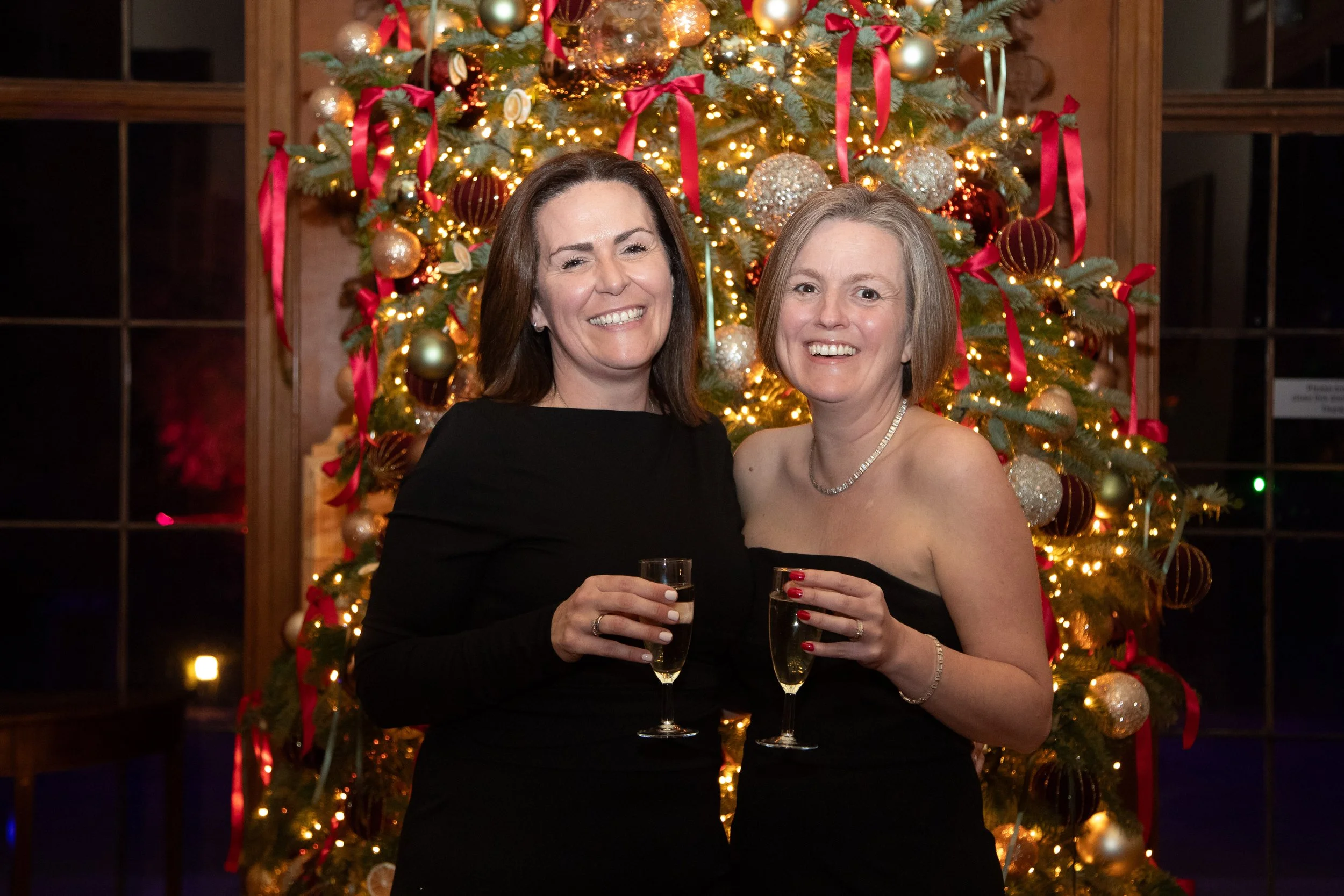 Two women in elegant black dresses are smiling and holding glasses of champagne in front of a decorated Christmas tree.