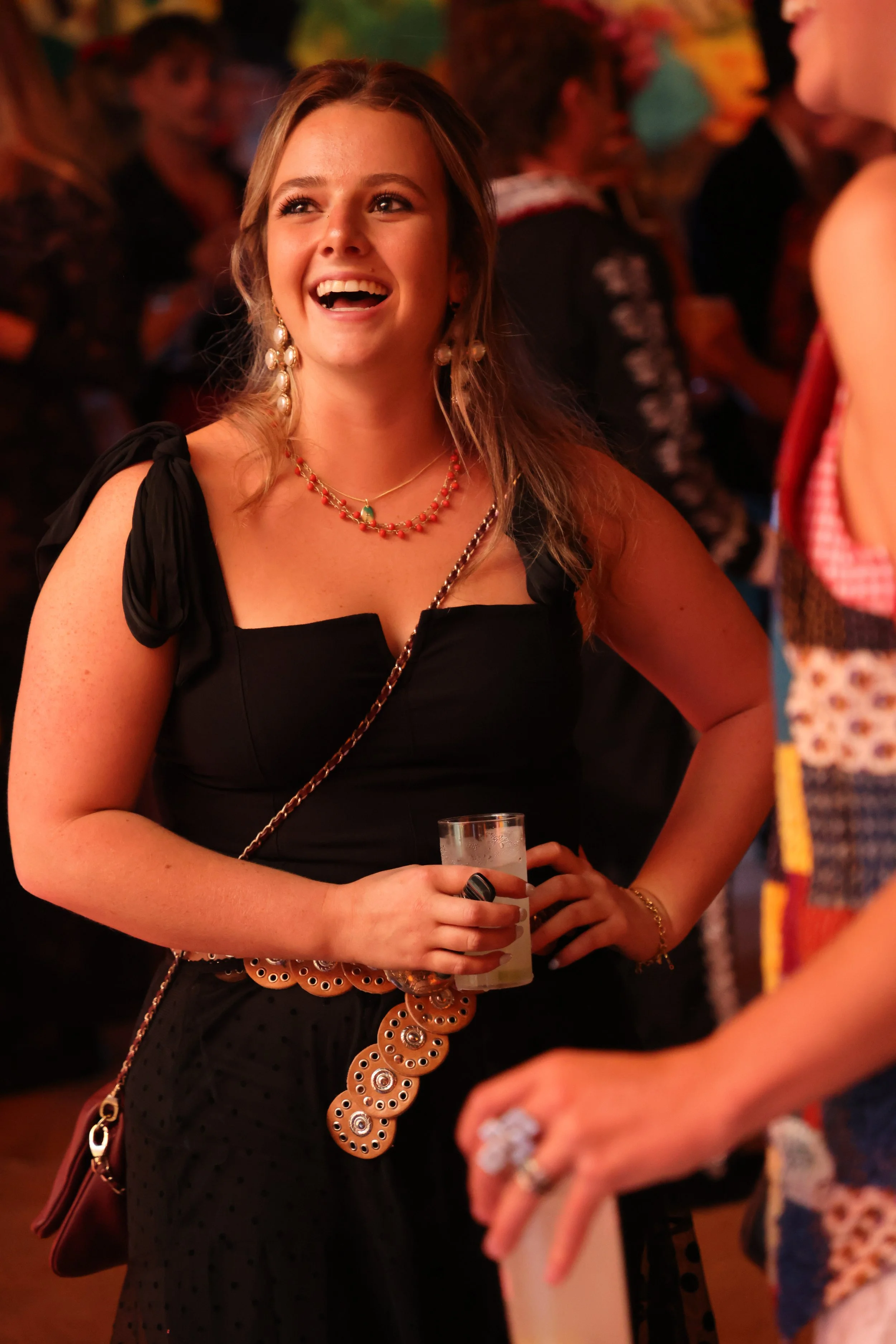 A young woman with long brown hair and earrings smiling, holding a glass of drink at a social event.