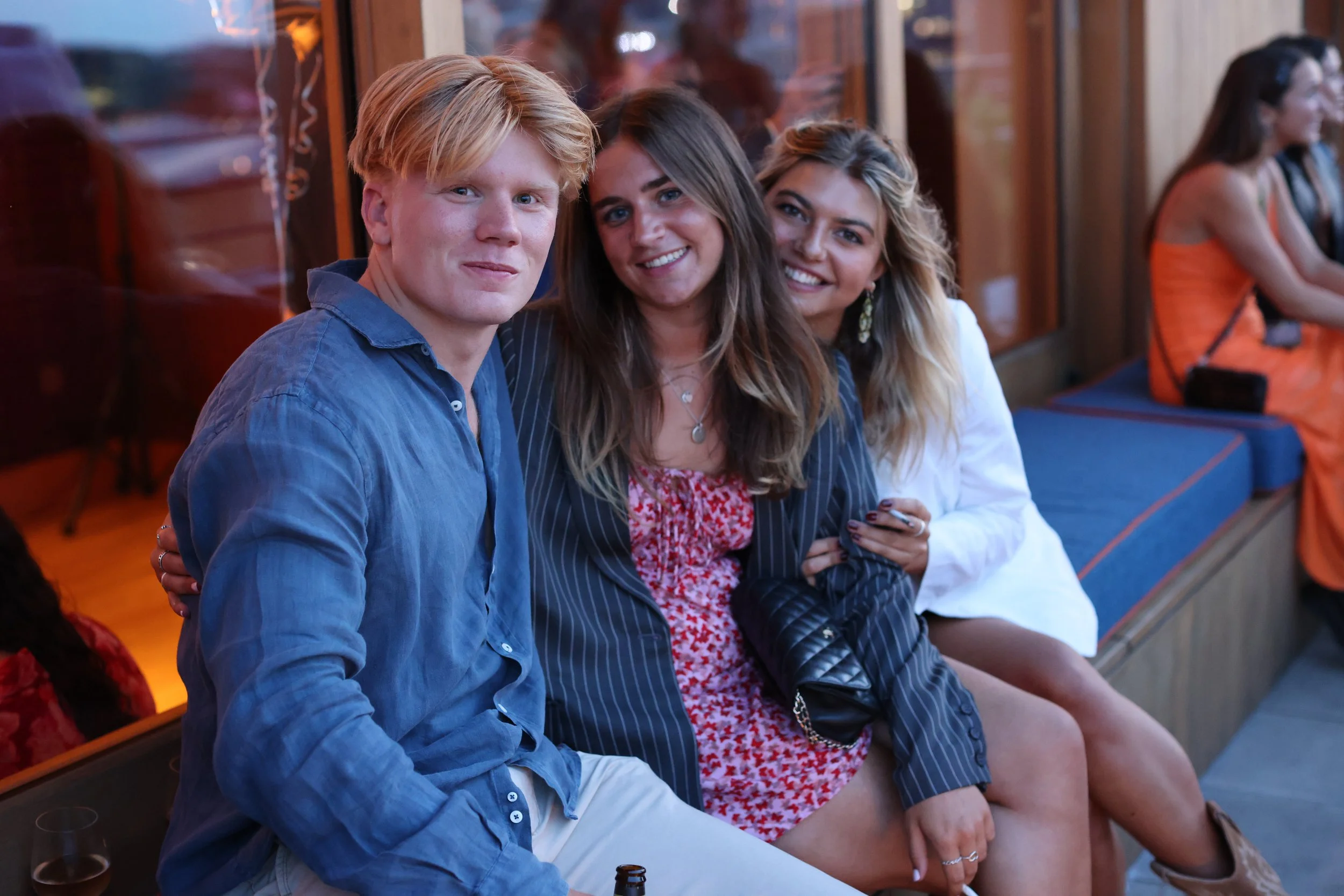 Four young adults sitting in a row at a social gathering, smiling and looking at the camera, with a woman in an orange top in the background.