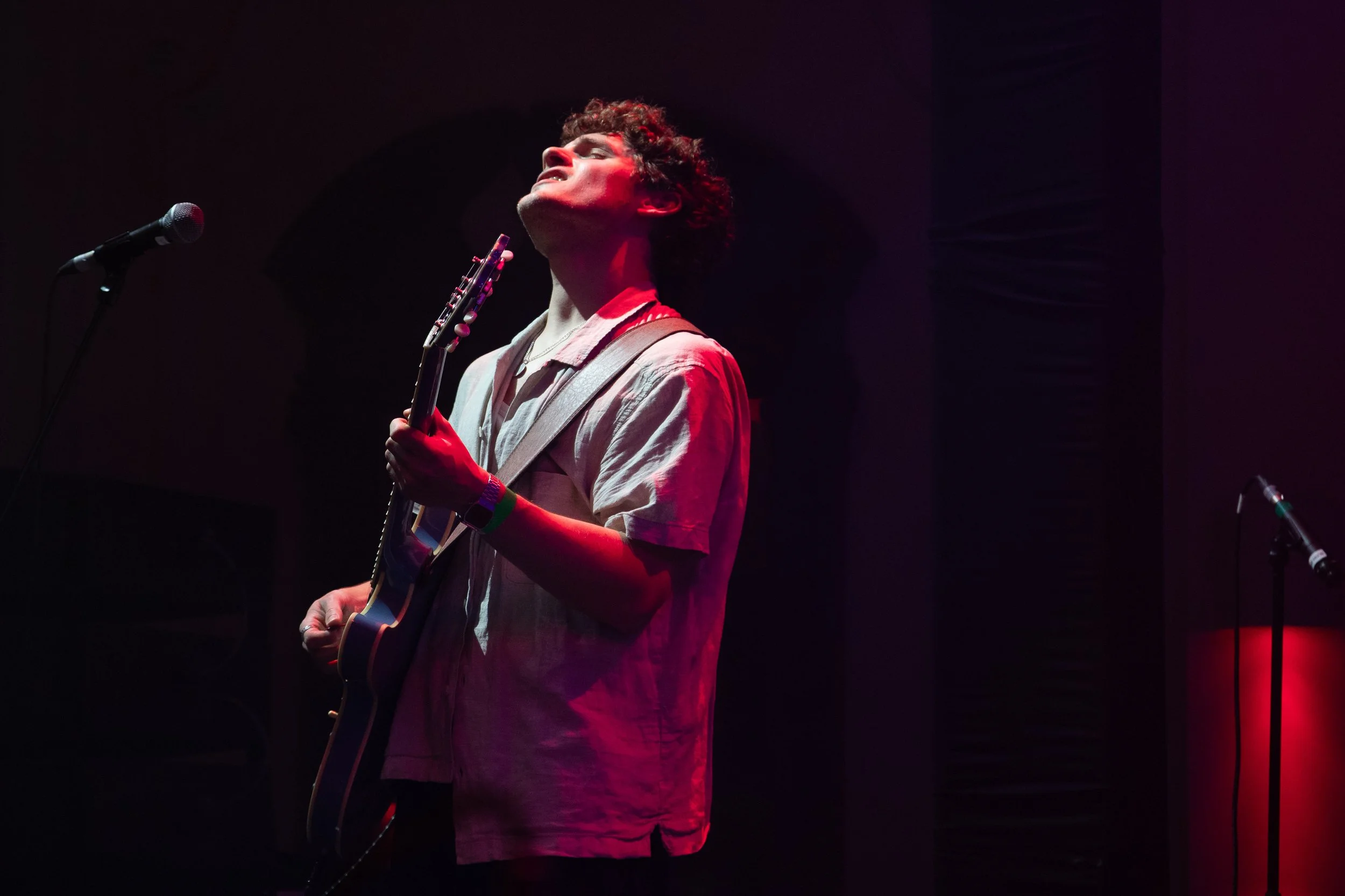 A person with curly hair singing passionately into a microphone on stage while holding an electric guitar, with a dark background and red stage lighting.