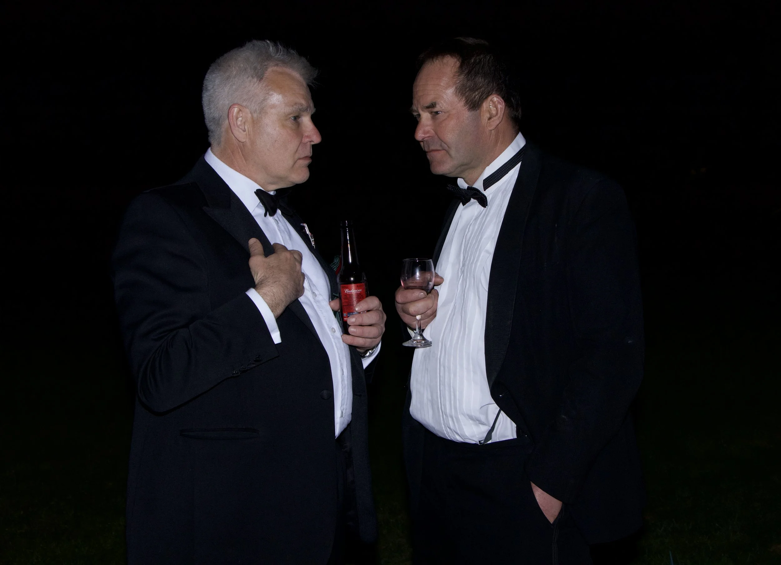 Two men in tuxedos face each other in an intense standoff at night, with one man holding a beer and the other holding a glass of wine.