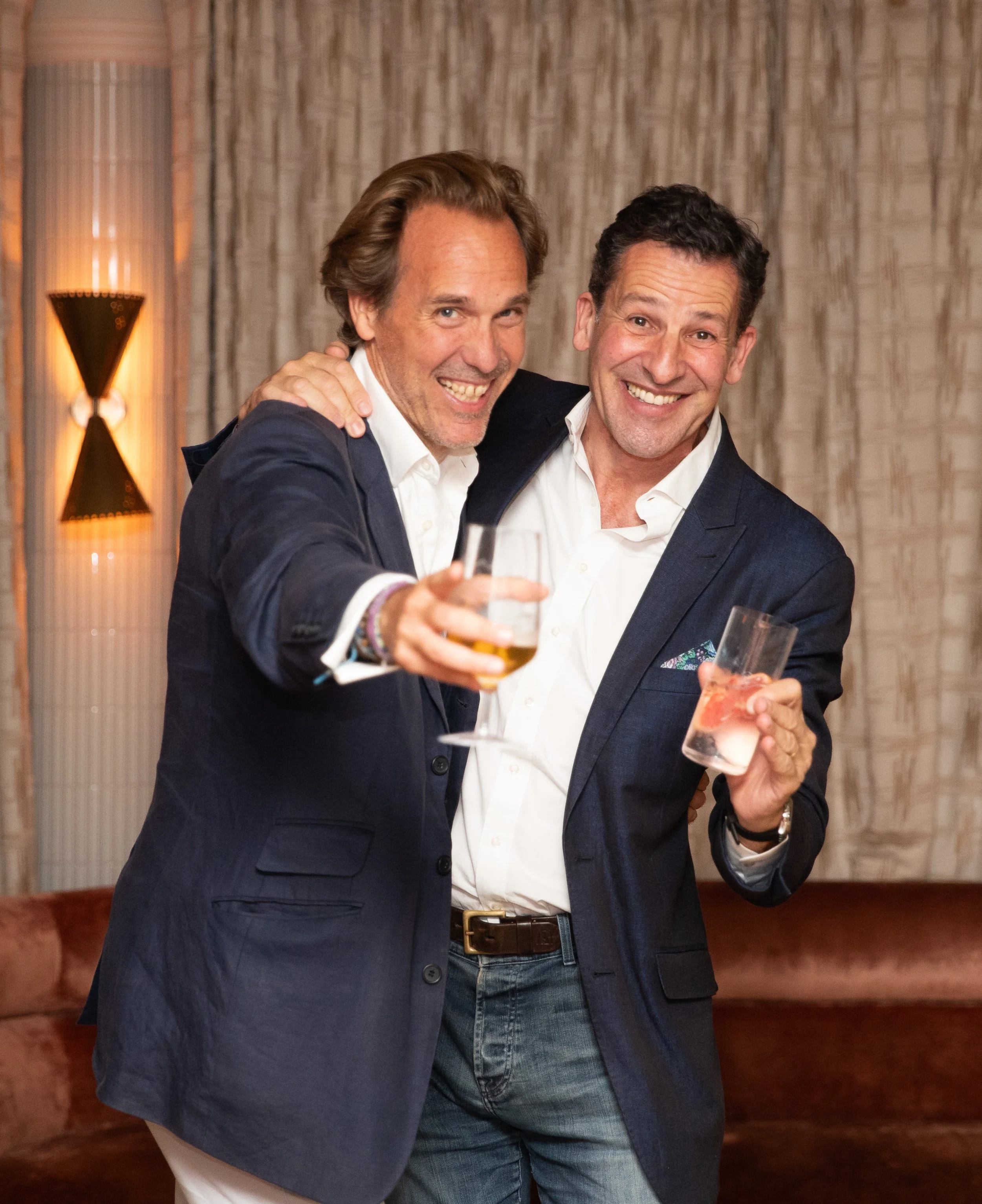Two men in suits smiling and raising glasses in a toast at a social gathering indoors.