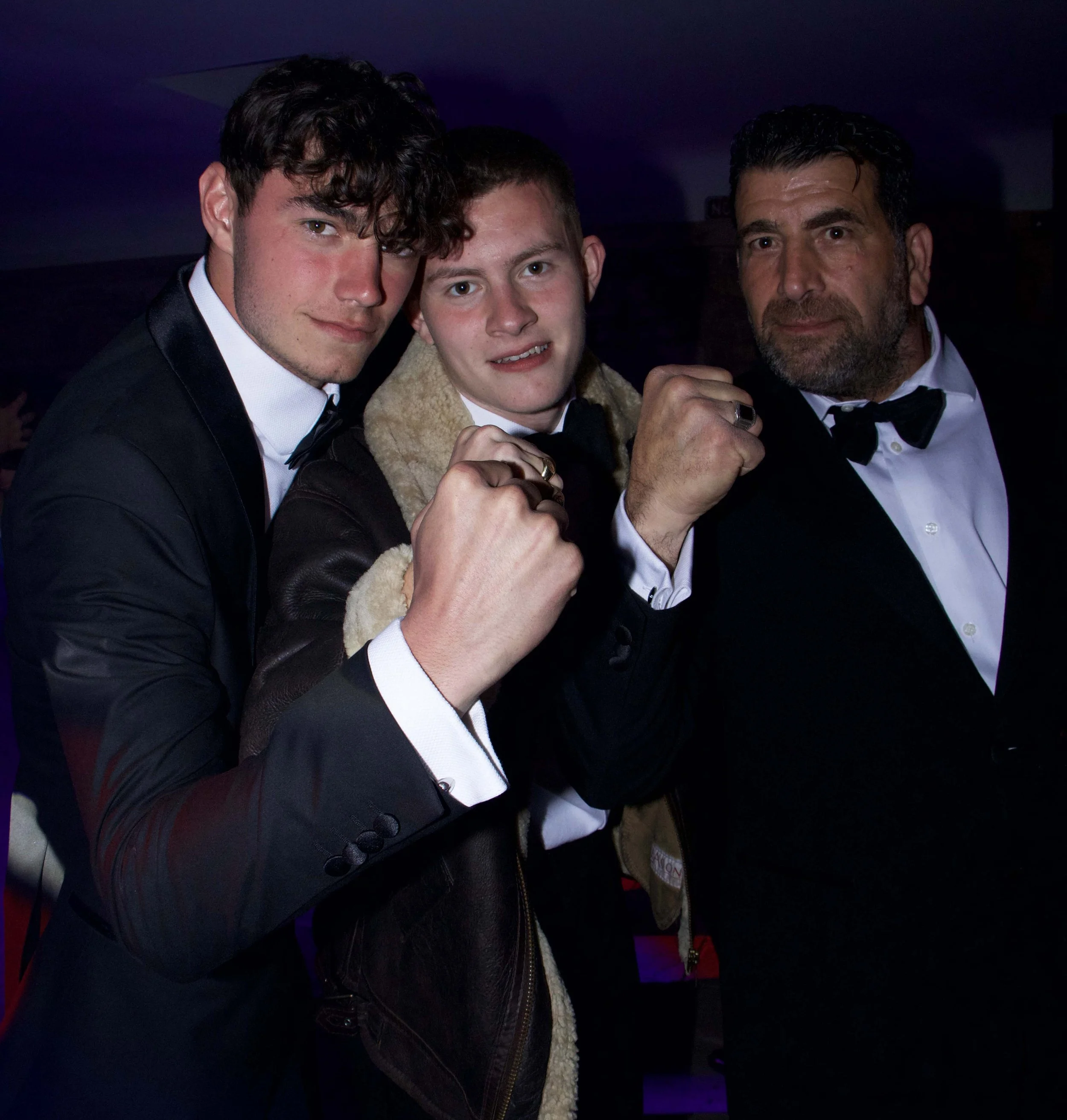 Three men in tuxedos with bow ties, two with fists raised, posing for a photo at an event, with dark background and purple lighting.