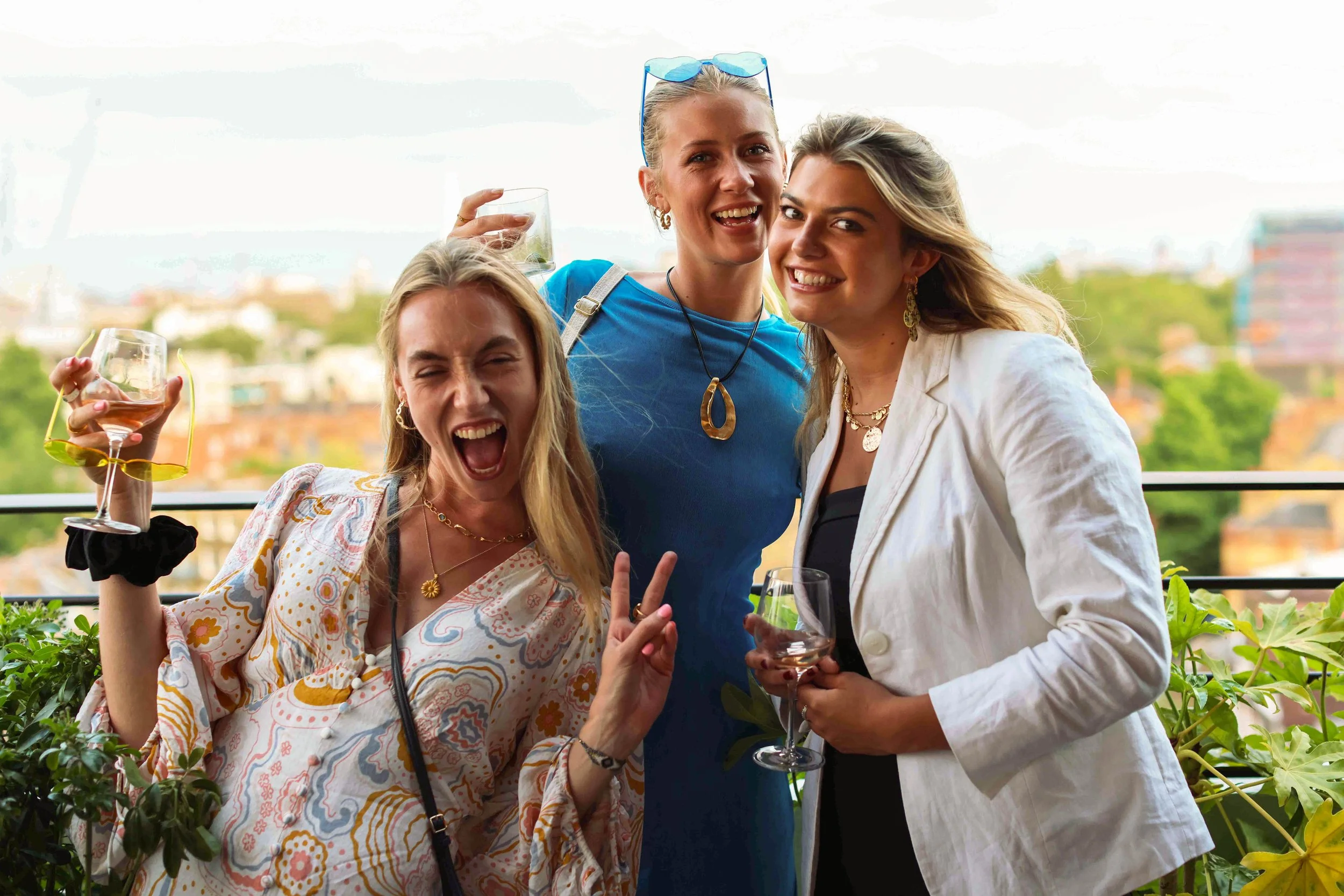 Three women at a social gathering, smiling and holding wine glasses, on a balcony with a city view in the background.