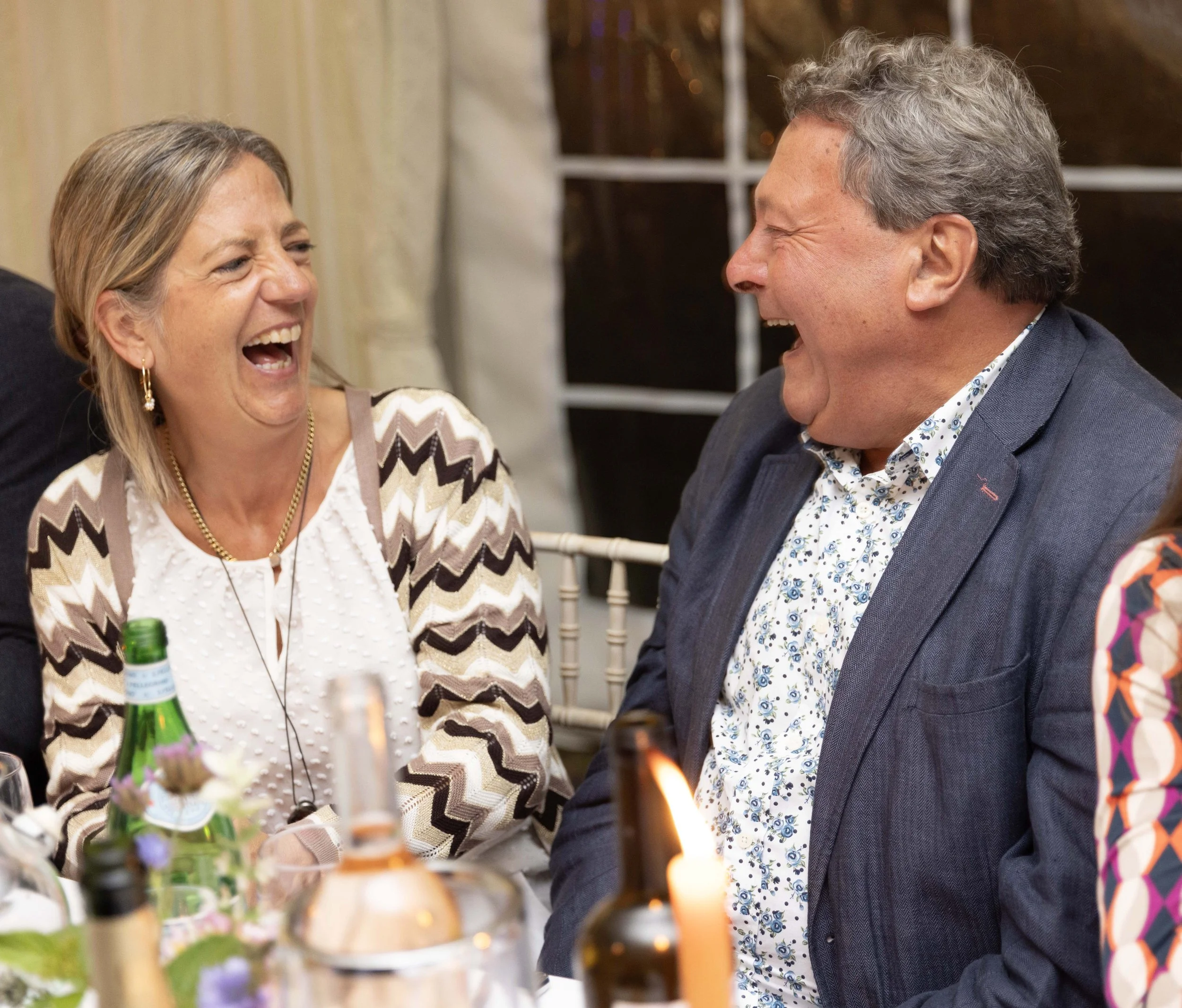 A woman and a man laughing and talking at a social gathering, surrounded by decorations and drinks.
