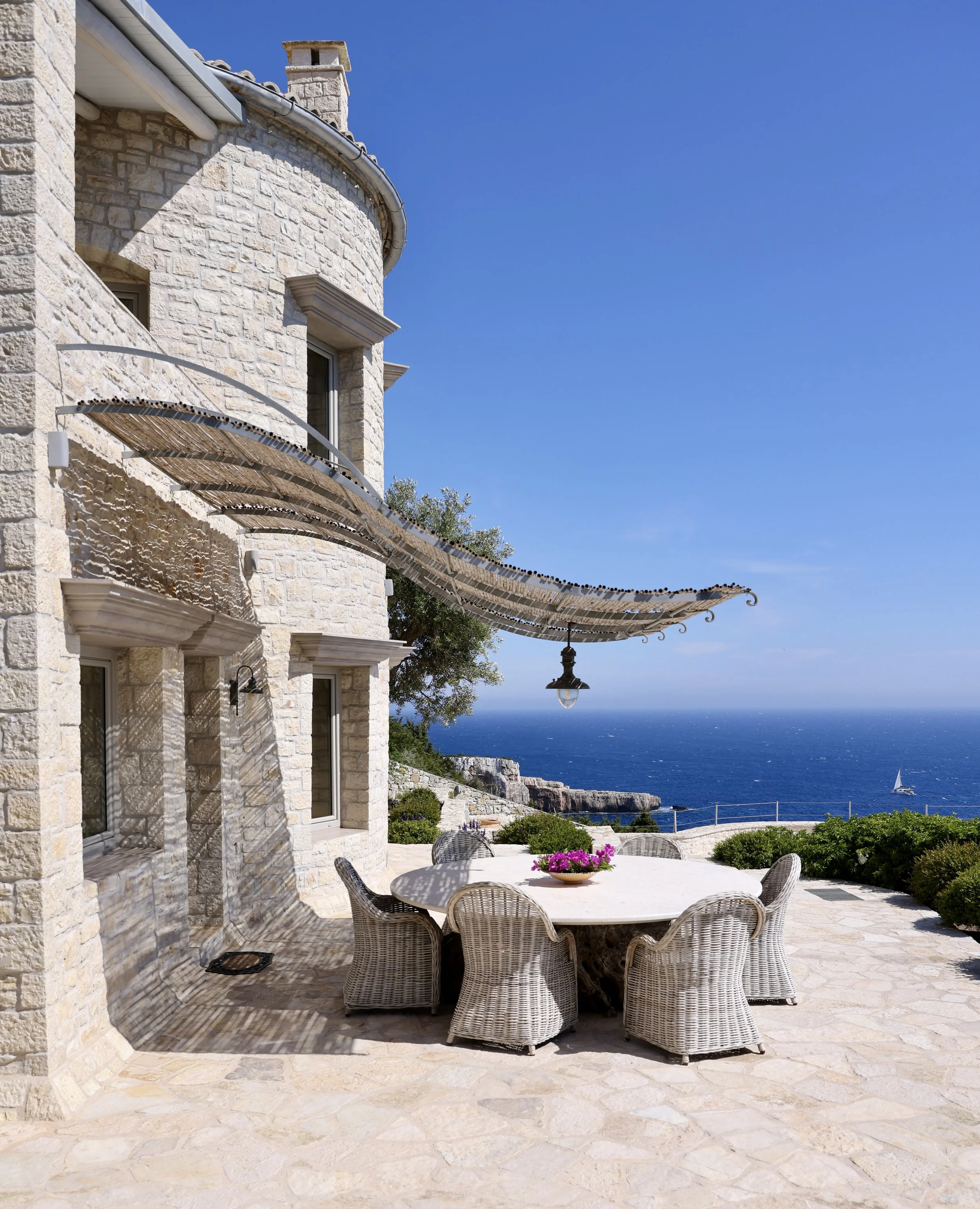 Stone house with outdoor patio overlooking ocean, round white table with wicker chairs, pink flowers in a bowl, blue sky, sailboat in water.