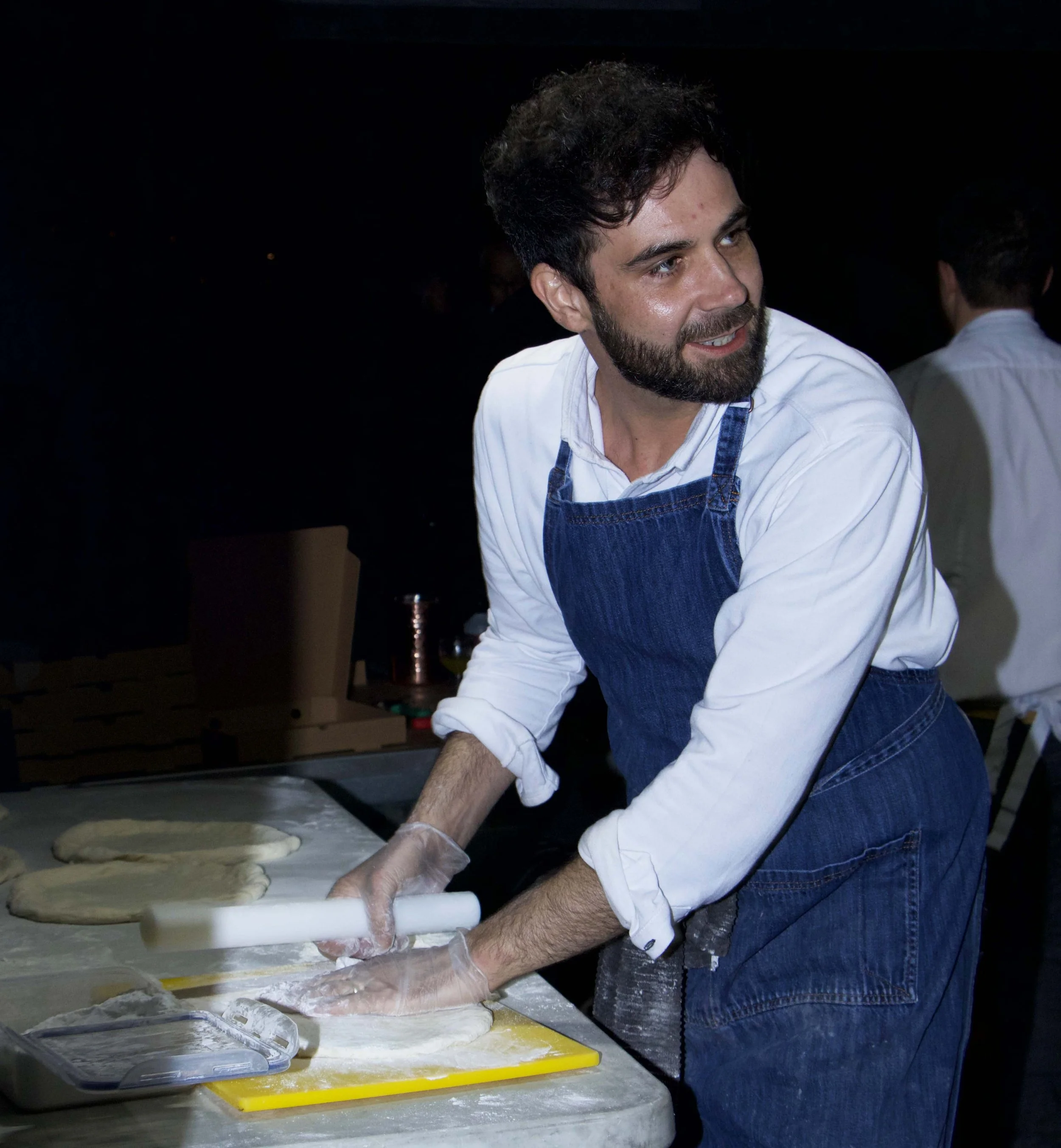 Man wearing a white shirt and blue apron rolling out dough on a yellow cutting board in a kitchen with a black background.