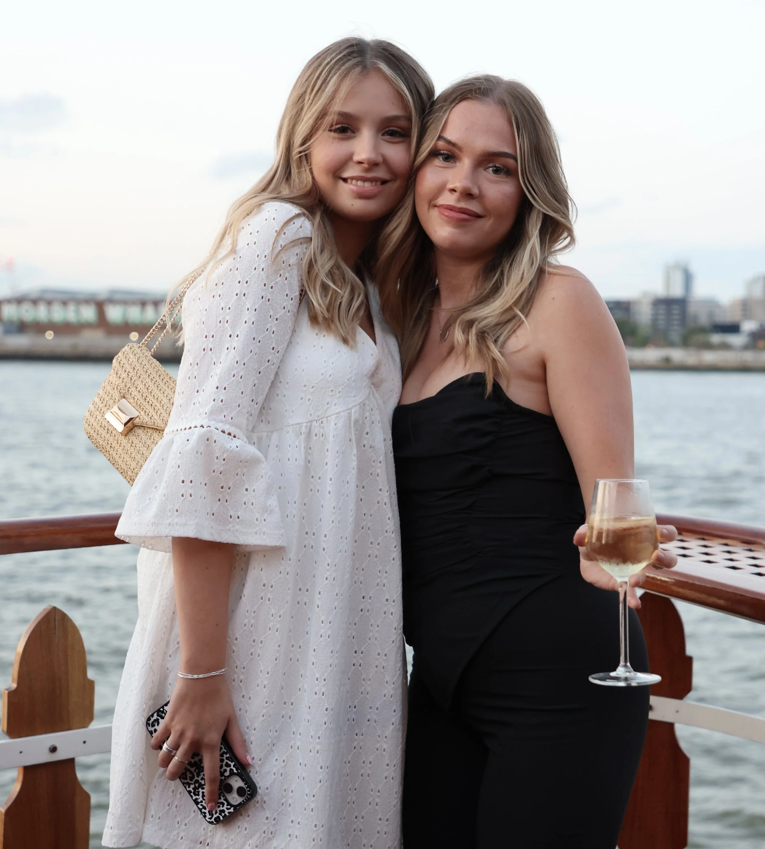 Two young women with blonde hair smiling on a boat, one in a white dress holding a phone with a leopard print case, the other in a black strapless dress holding a glass of white wine, with a city skyline and water in the background during the evening