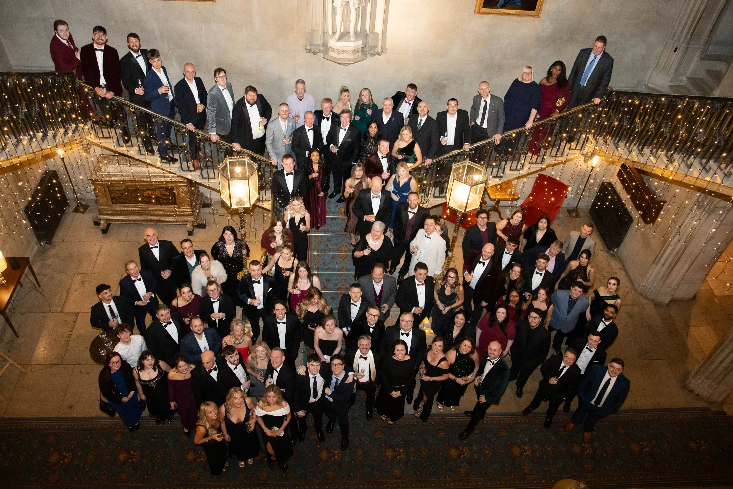 A large group of people attending a formal event in a grand hall, standing on a decorated staircase, dressed in tuxedos and evening gowns, holding drinks, with a pipe organ and religious statues visible in the background.