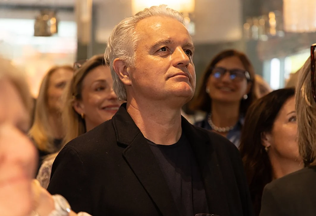 An older man with silver hair and a serious expression at a social event, surrounded by women smiling and looking in the same direction.