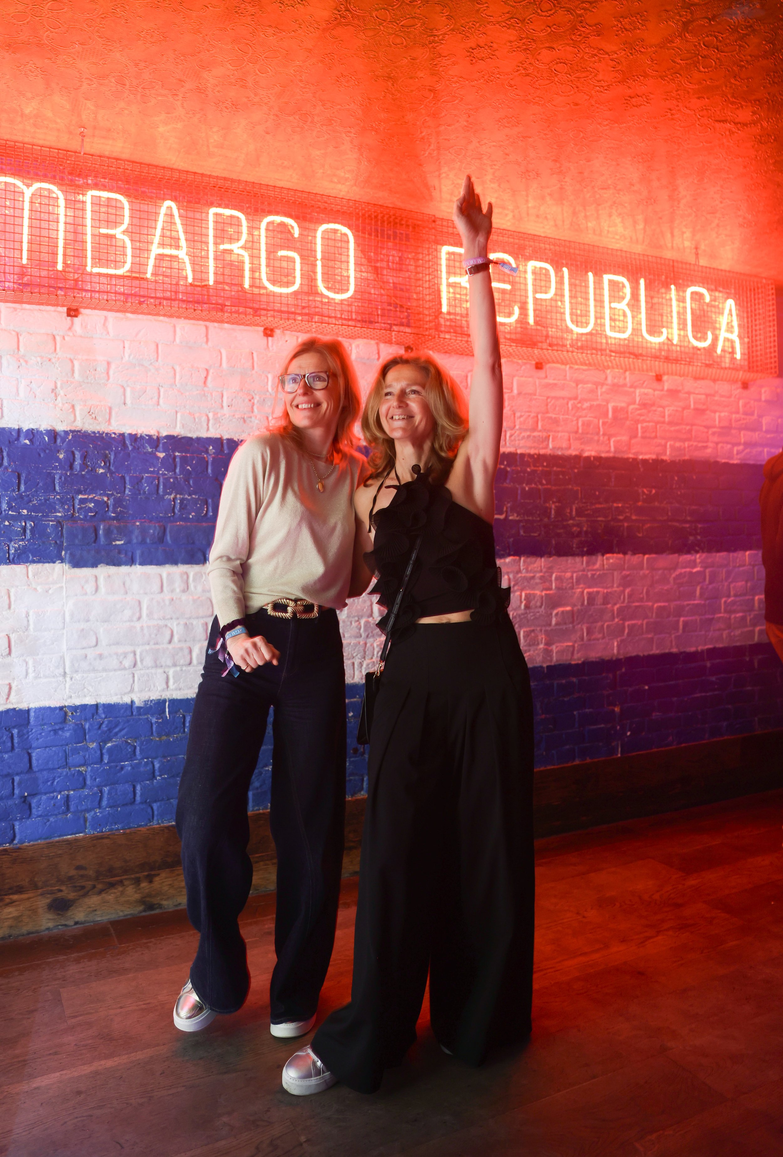 Two women standing in front of a neon sign that says 'Embargo Republica'; one with glasses and a beige sweater, the other with light hair and a black top raising her right arm. The background includes a brick wall painted with red, white, and blue st