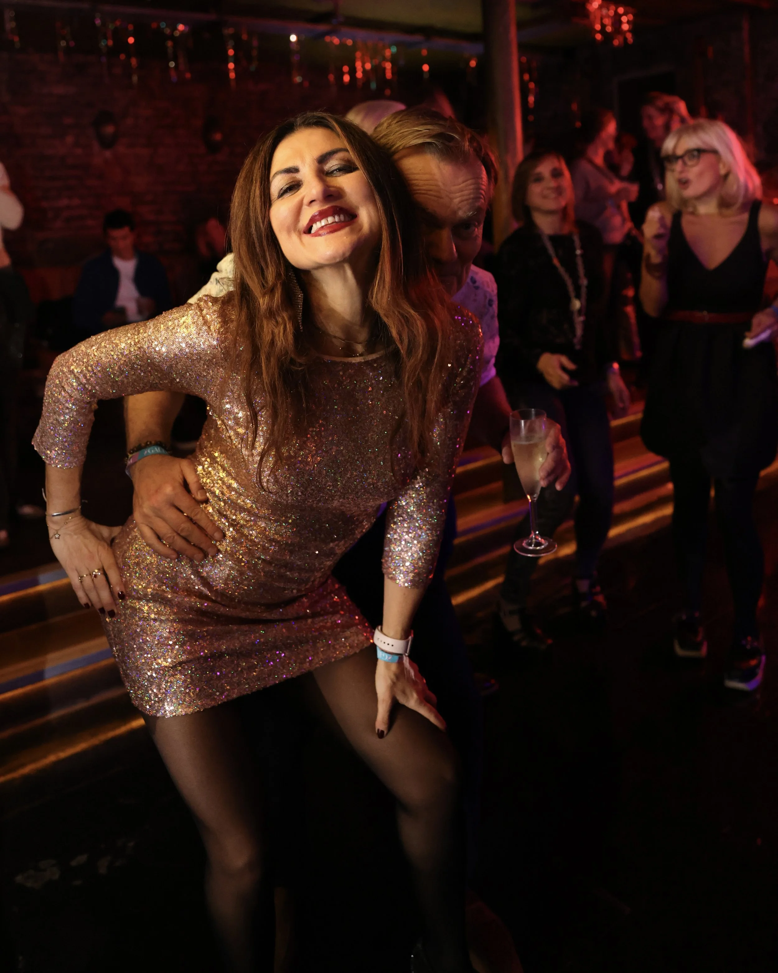 Woman in a glittery gold dress smiling and posing with a hand on her hip at a party or nightclub, surrounded by other people dancing and socializing in a dimly lit room with red lighting and brick walls.