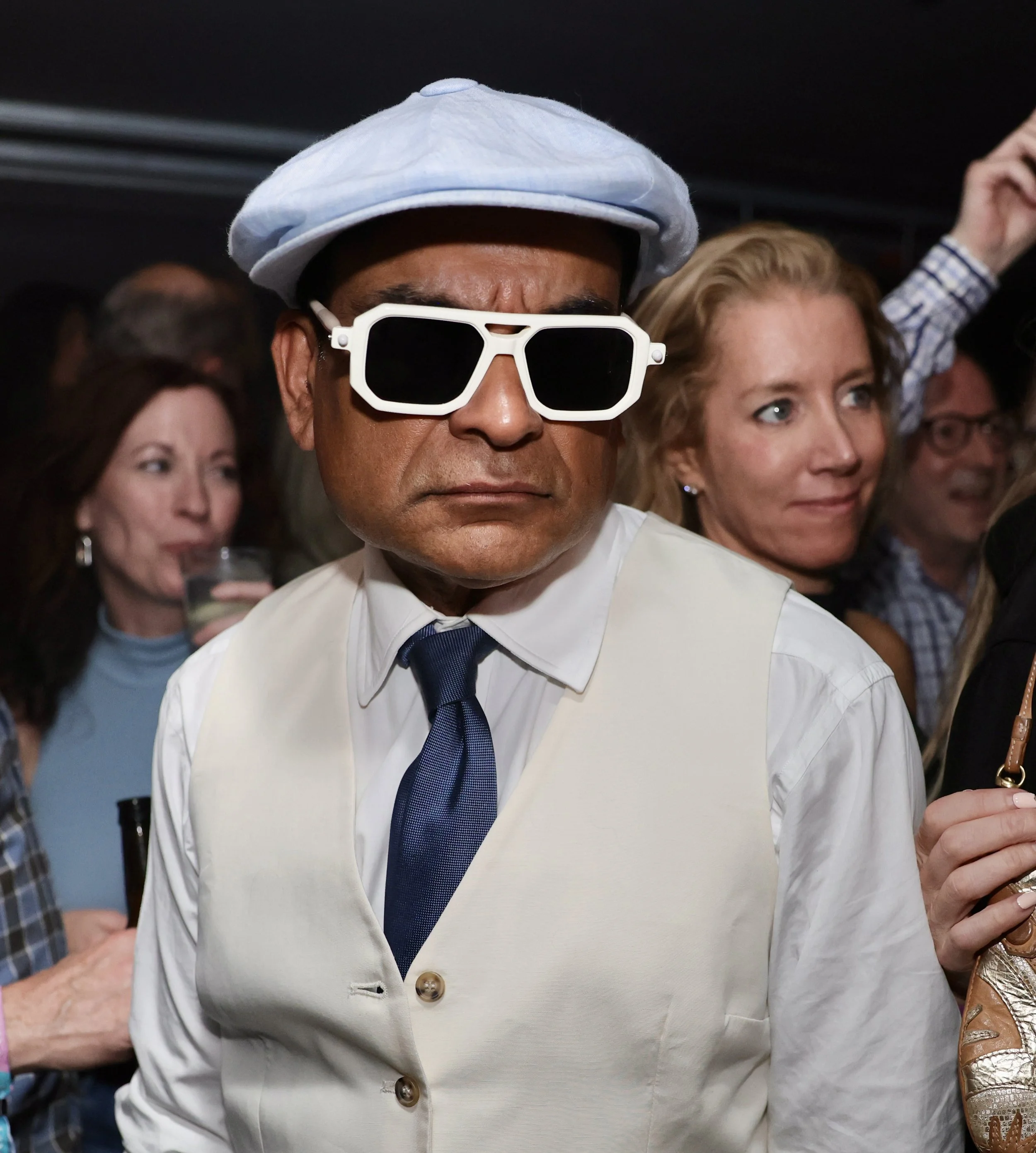 A man wearing a white cap, large white sunglasses, a white vest, and a blue tie at a social event.