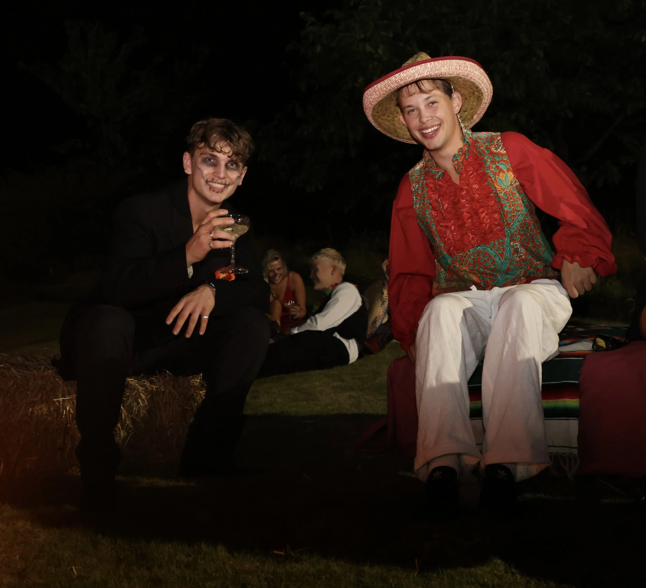 Two young men at an outdoor nighttime gathering. The man on the left is dressed in dark clothing with Halloween makeup, holding a cocktail glass, and smiling. The man on the right is dressed in bright, colorful clothing with a large straw hat, also s