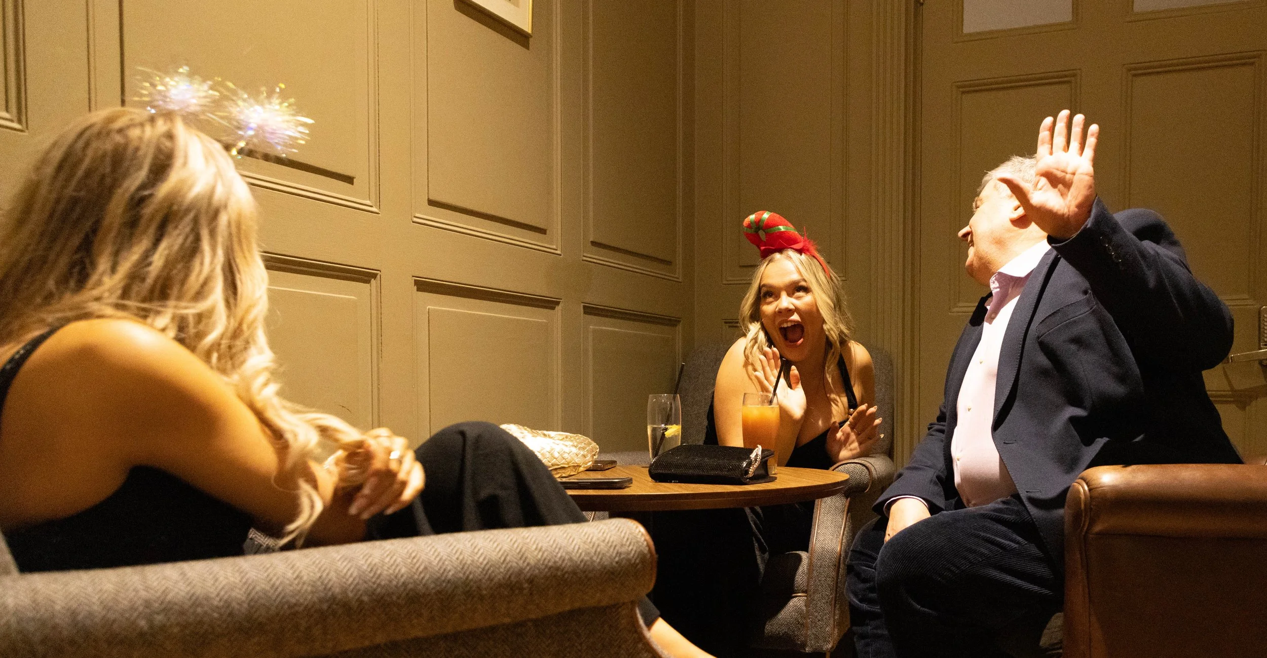 Four people sitting around a table at a social gathering, two women and two men, with holiday hats and accessories, engaged in conversation and laughter in a warmly lit room.
