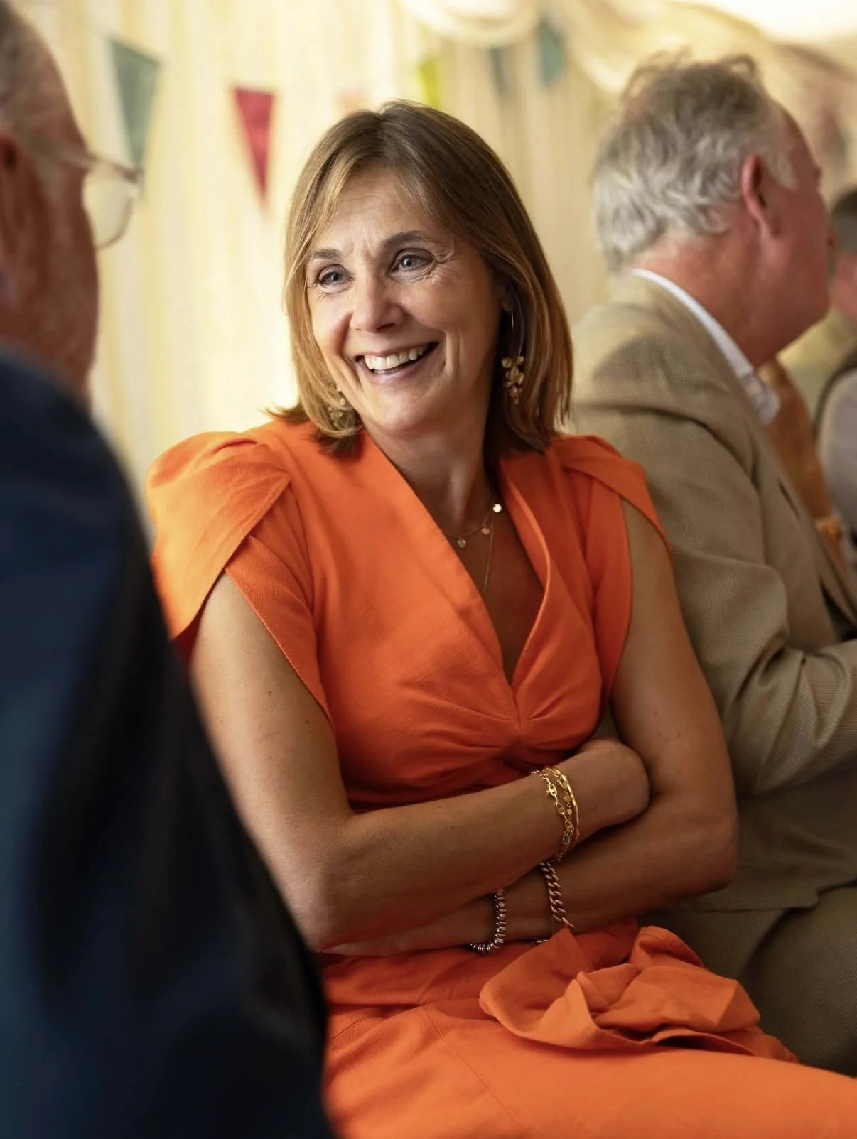 A woman in an orange dress is sitting at a table, smiling and engaging with others in a social gathering, with a man in a beige jacket beside her.
