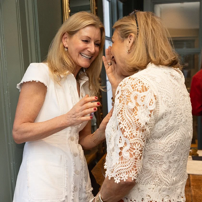 Two women with blonde hair, dressed in white, smiling and whispering to each other in an indoor setting.