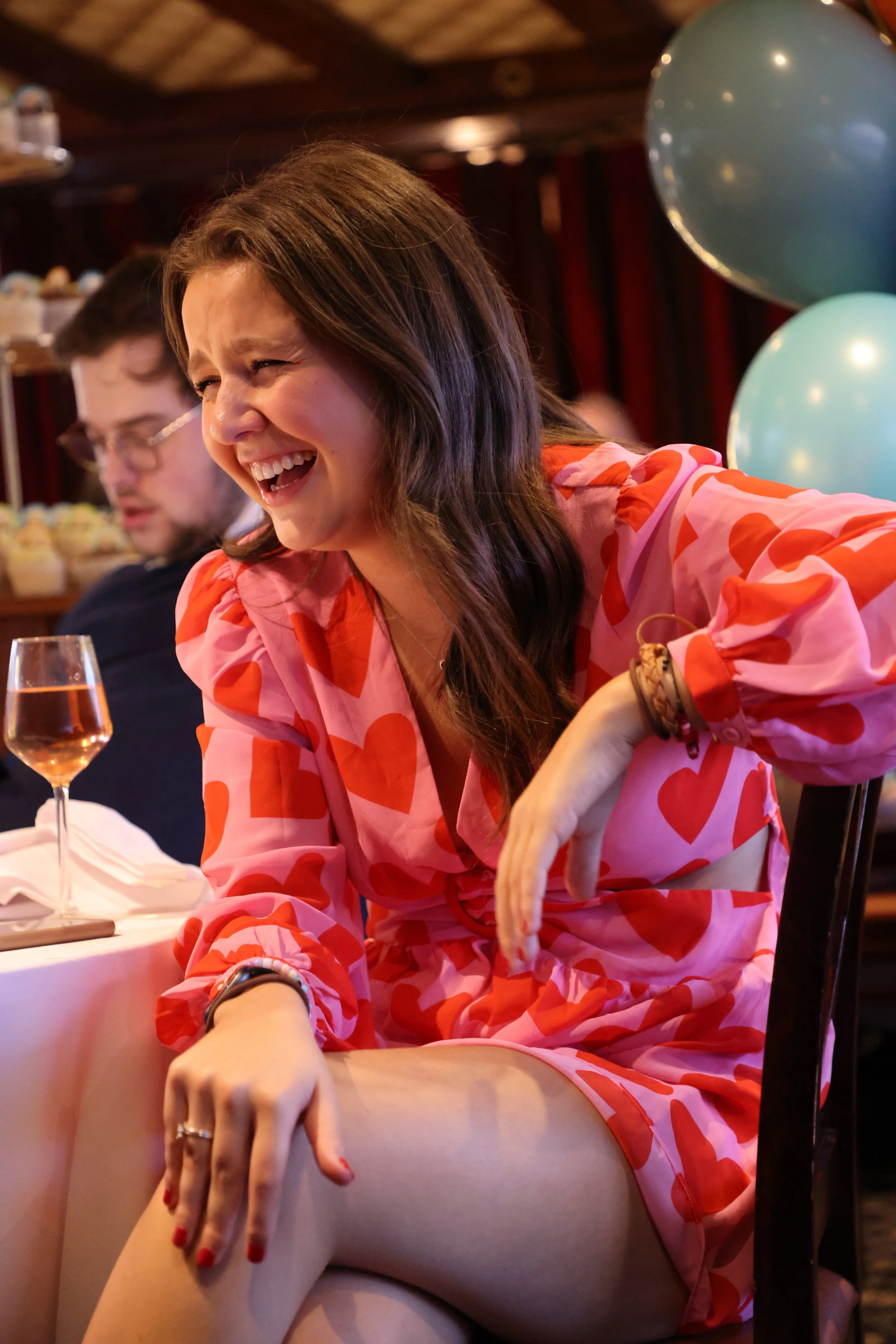 A woman in a pink dress with red hearts laughing at a celebration or party, sitting at a table with balloons and a glass of rosé wine.