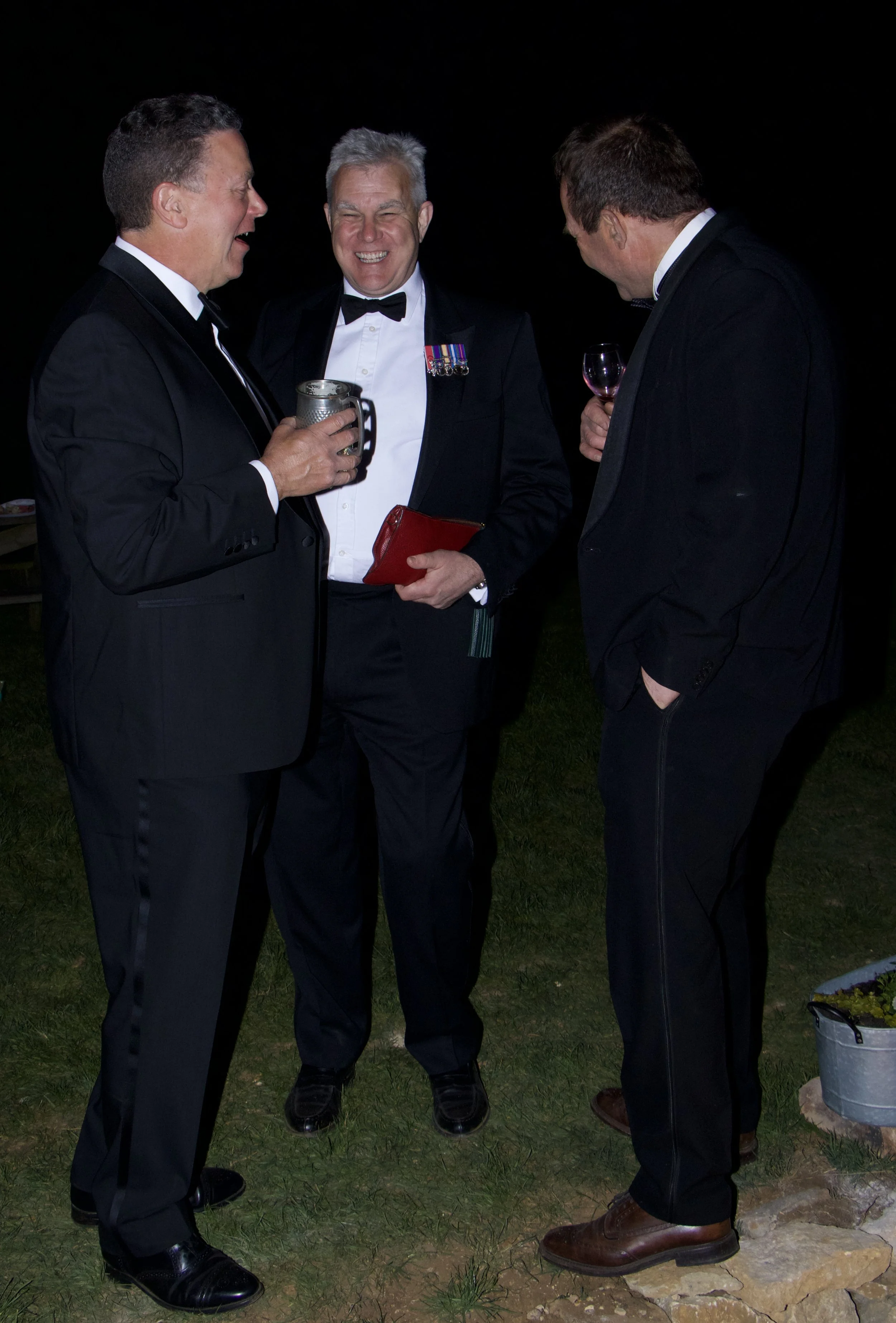 Three men in tuxedos laughing and talking outdoors at night, holding drinks.