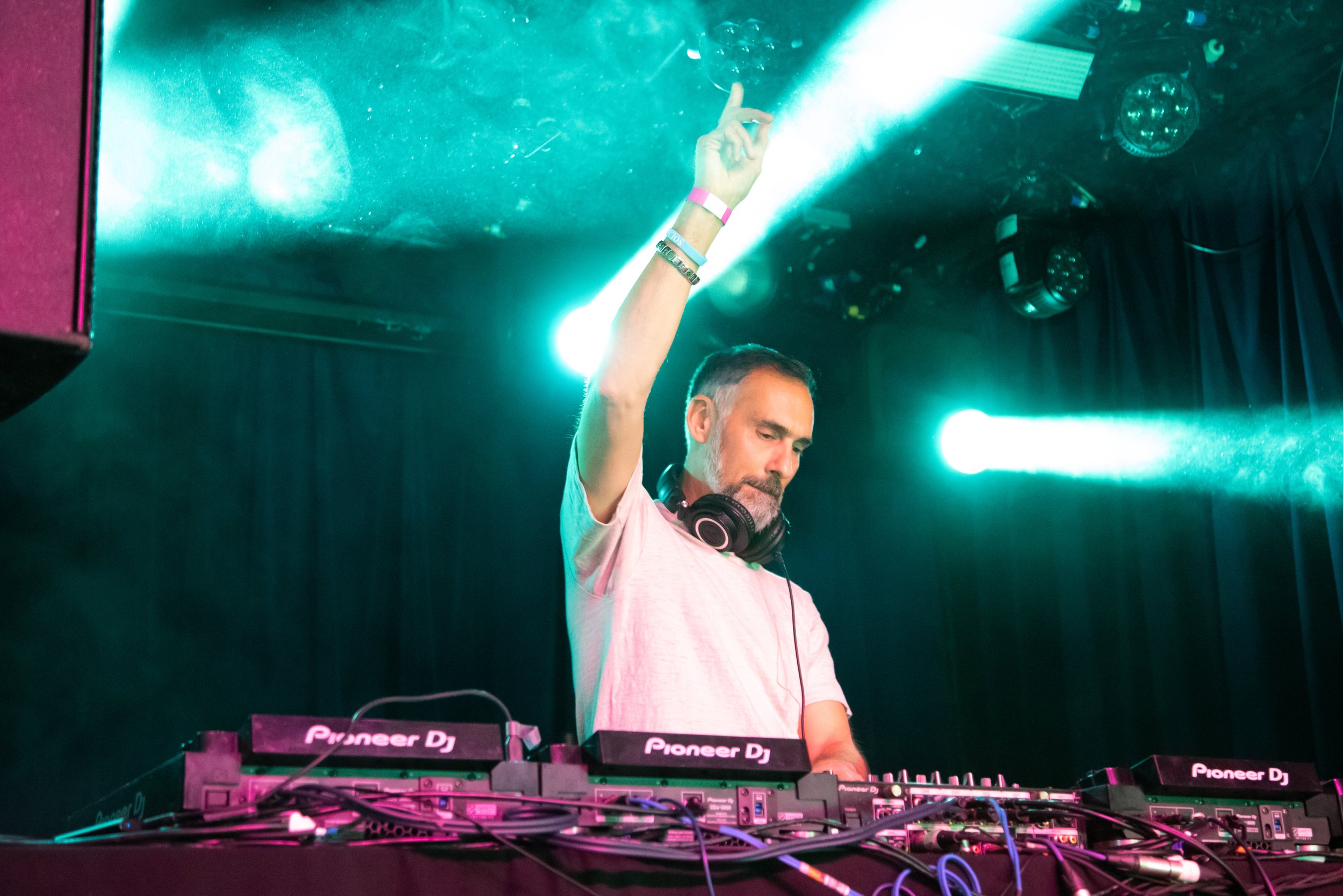 A DJ with a beard and short hair, wearing headphones around his neck and a pink T-shirt, is in a nightclub or concert setting. He is raising his left hand with his index finger pointing up. There are Pioneer DJ equipment and colorful lighting around 