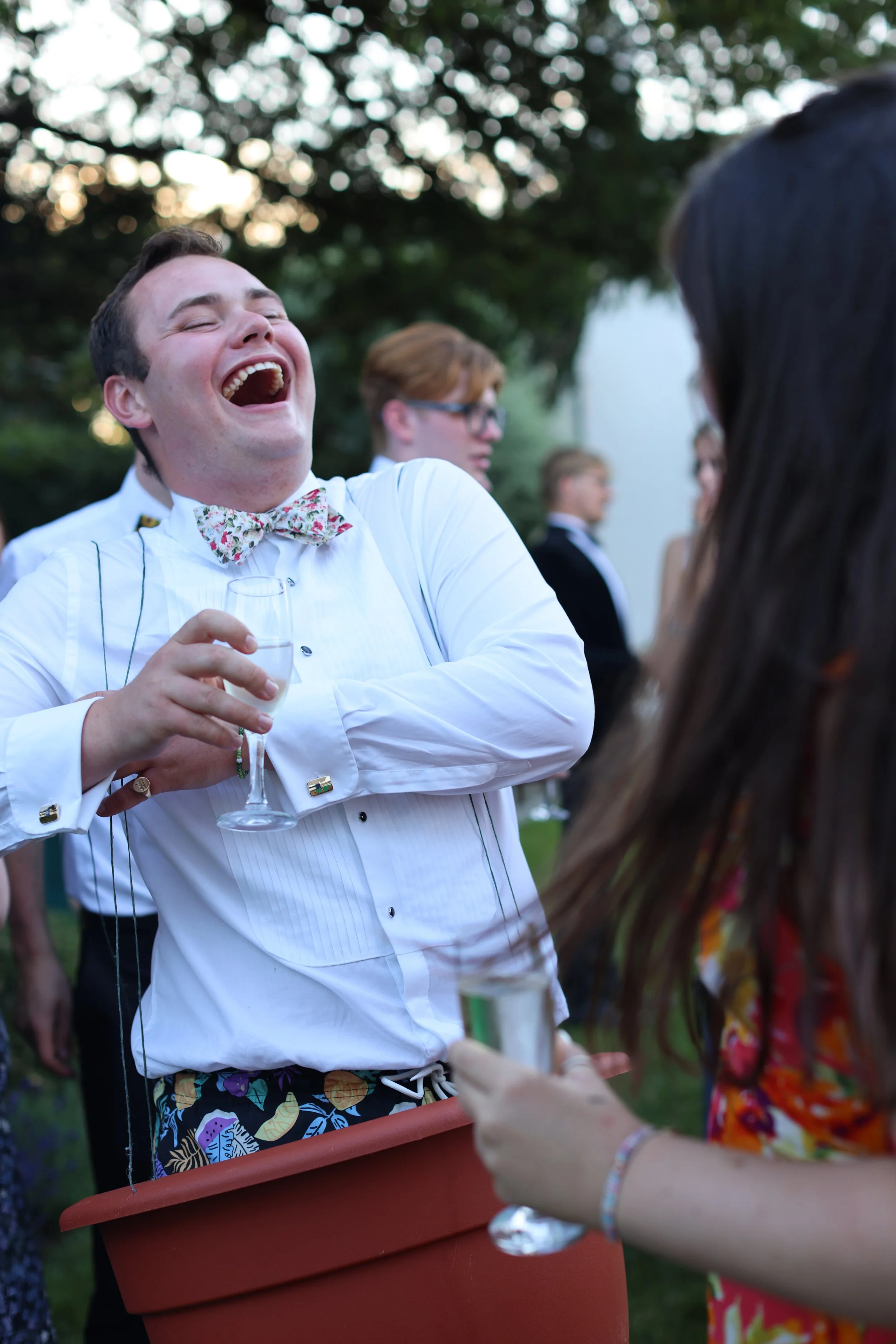 A man wearing a white shirt with a floral bow tie laughing while holding a glass of drink, standing near a woman with long dark hair in an outdoor setting with other people in the background.
