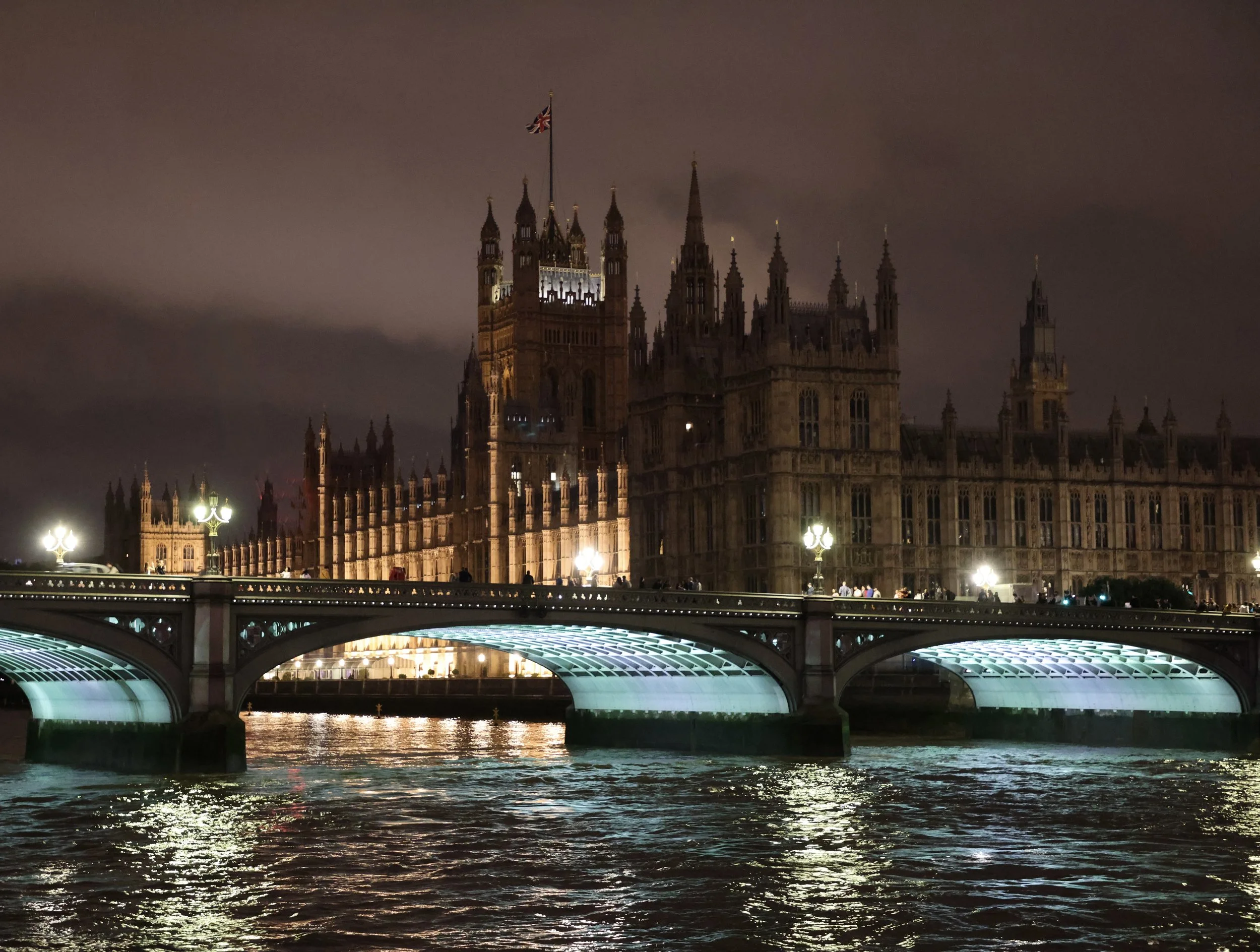 Night view of the Palace of Westminster in London, illuminated and reflected on the River Thames, with a bridge in the foreground.