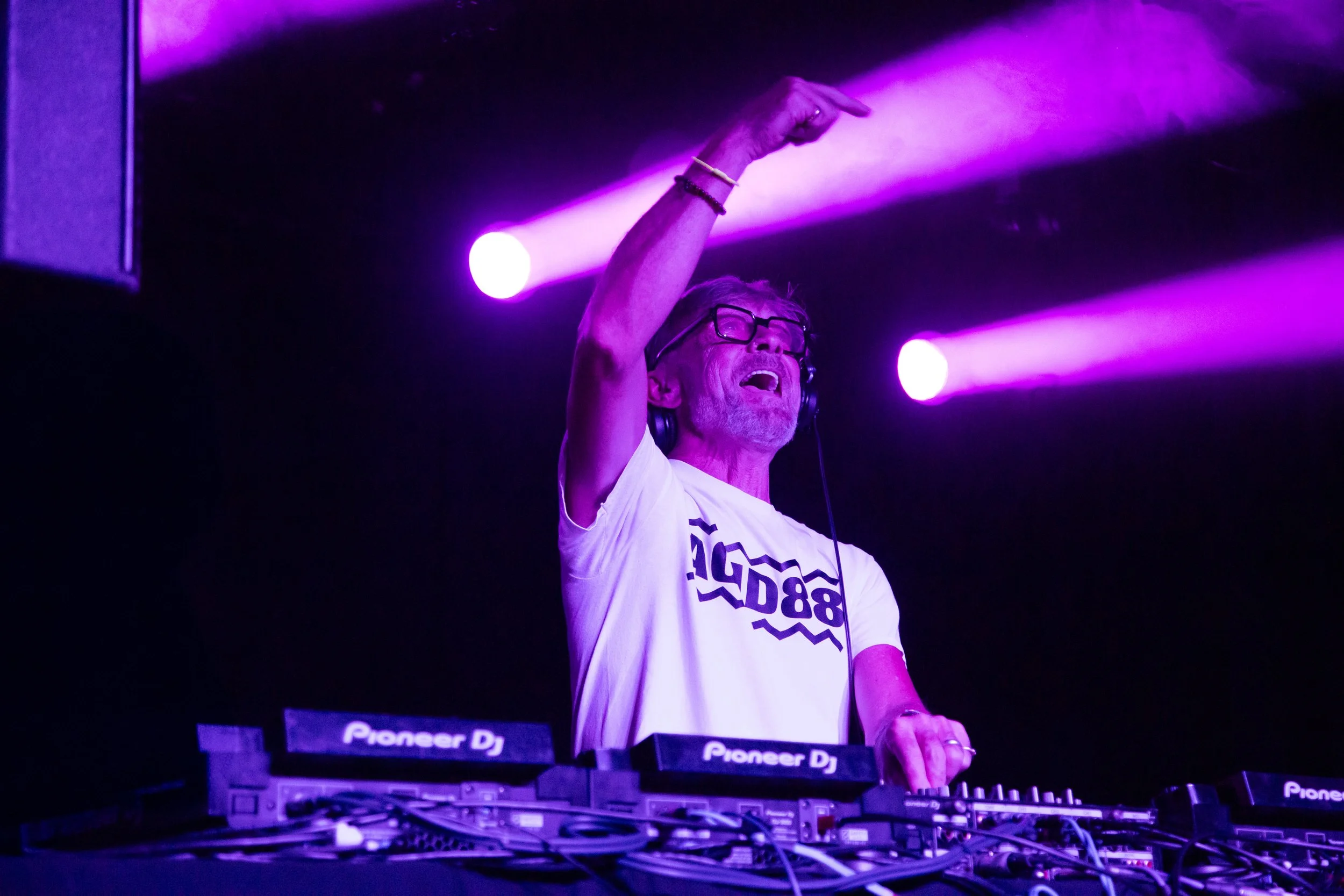 A DJ with glasses and a white T-shirt is performing at a dark stage illuminated with purple lights, standing behind a Pioneer DJ equipment setup, with one hand on the keys and the other hand raised.