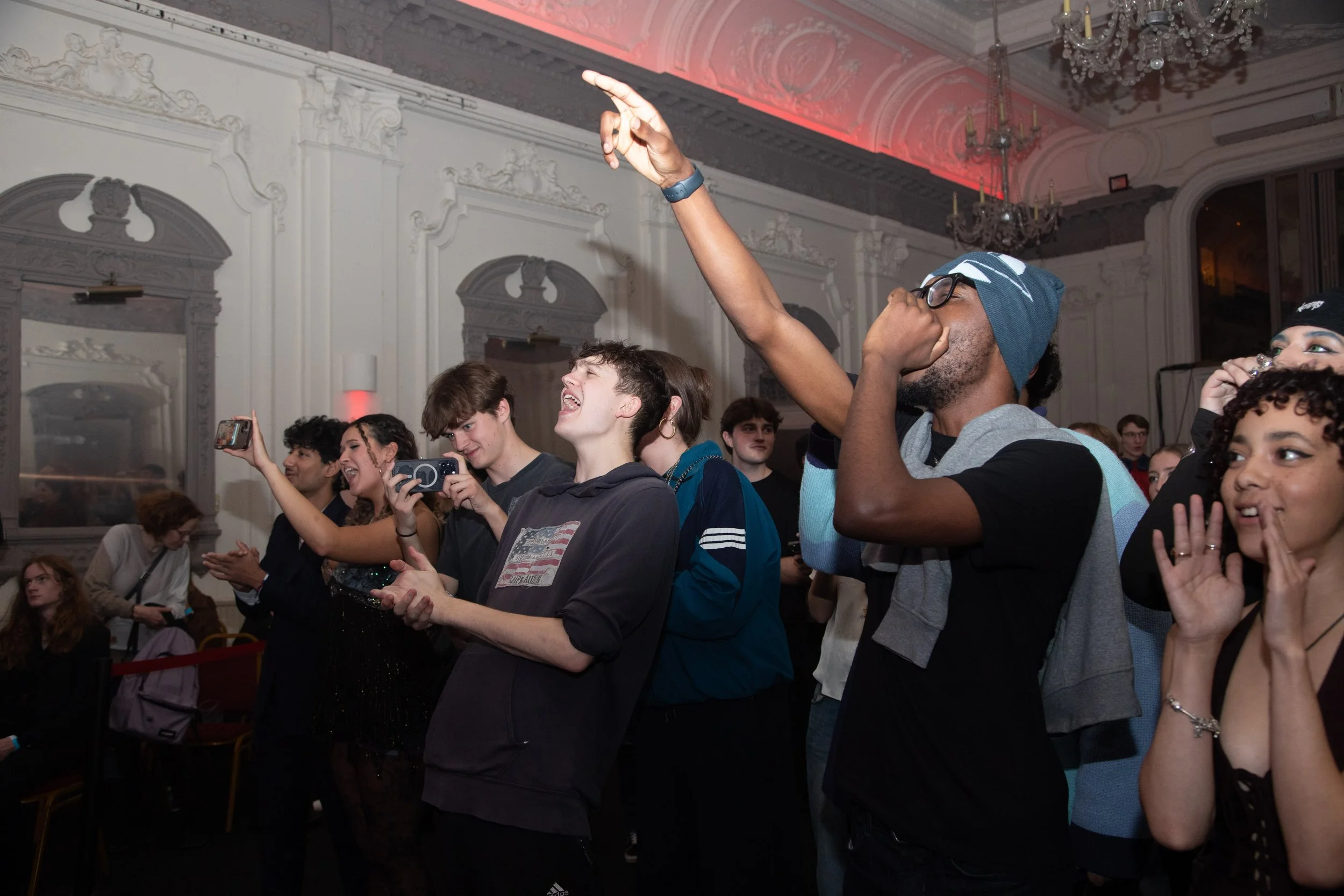 Group of people at an event, smiling, taking photos, and enjoying themselves in a decorated hall with chandeliers and ornate moldings.