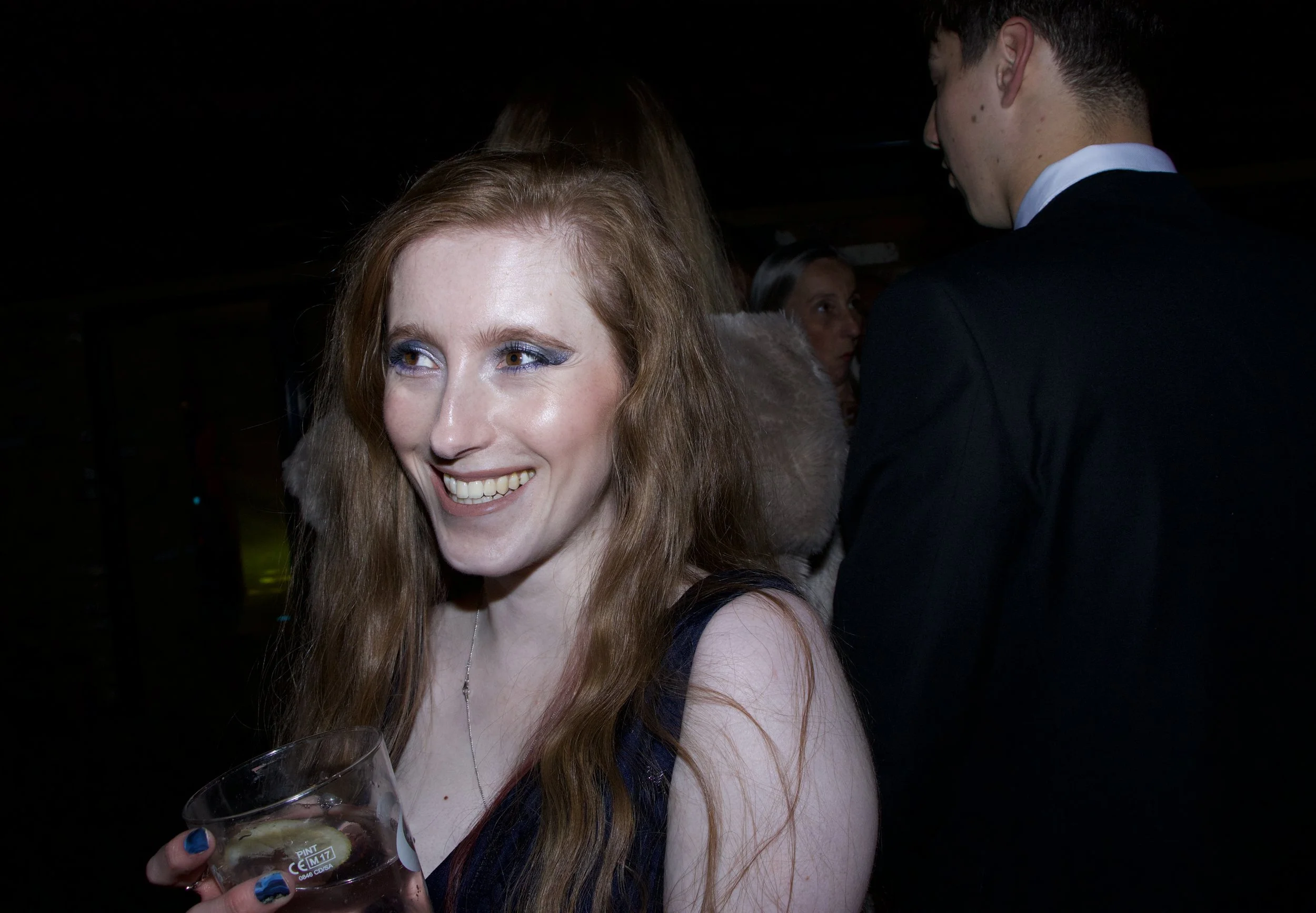 A woman with long red hair and blue eye makeup smiling, holding a glass of wine, with a man in a tuxedo in the background at a social event.