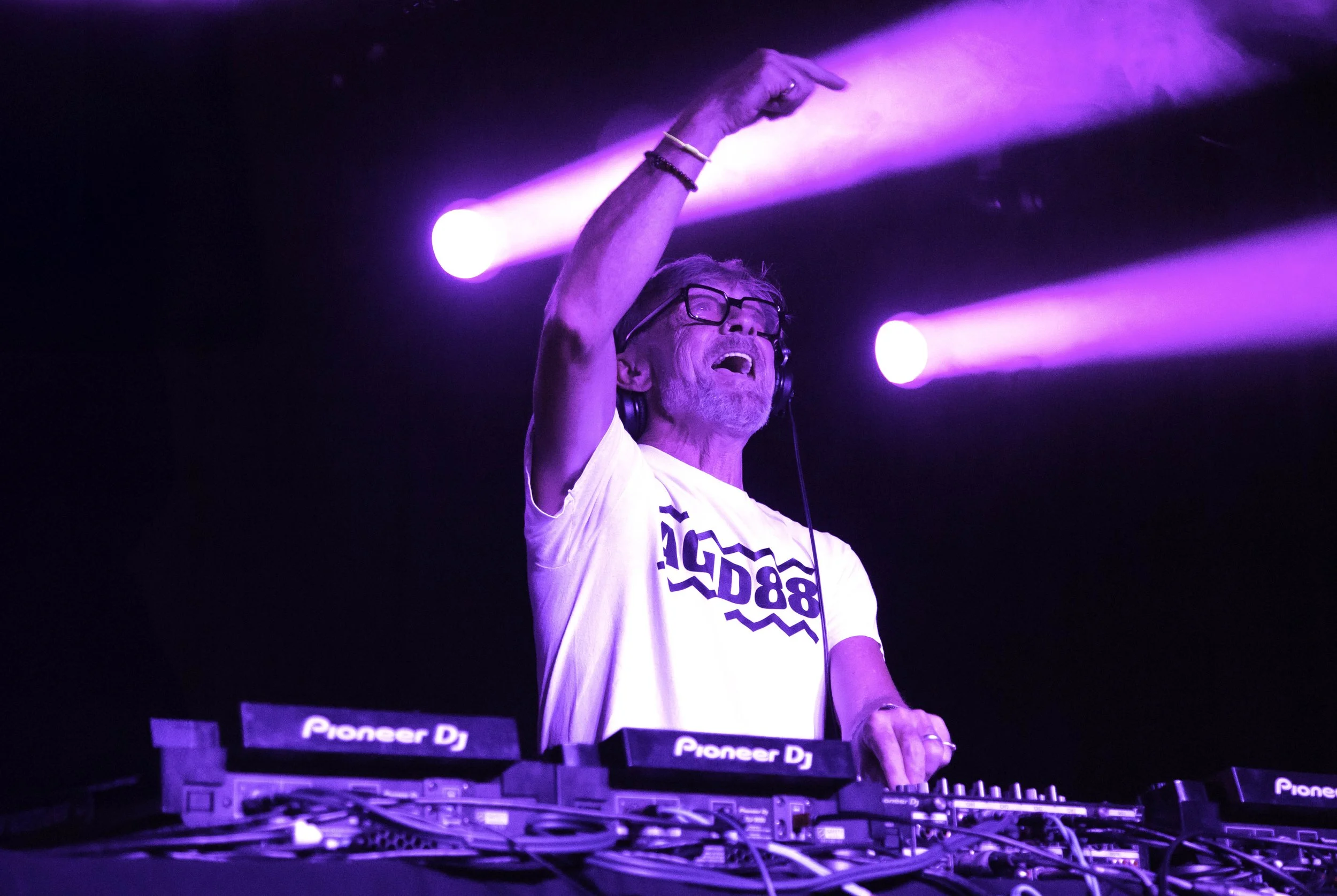 A DJ wearing glasses and a white T-shirt with black text, smiling and raising one arm, in a dark venue illuminated by purple stage lights, with DJ equipment labeled "Pioneer DJ" in front.