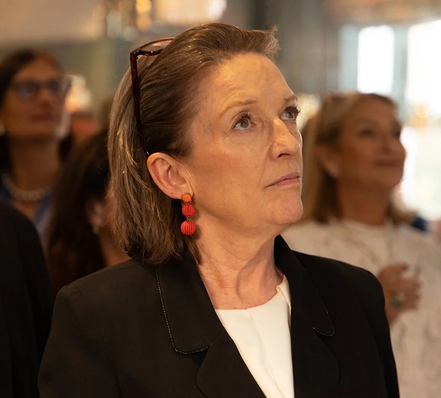 A woman with light skin, short brown hair, wearing red dangling earrings, a black blazer, and glasses on her head, looking attentively at something off-camera in a crowded indoor setting.