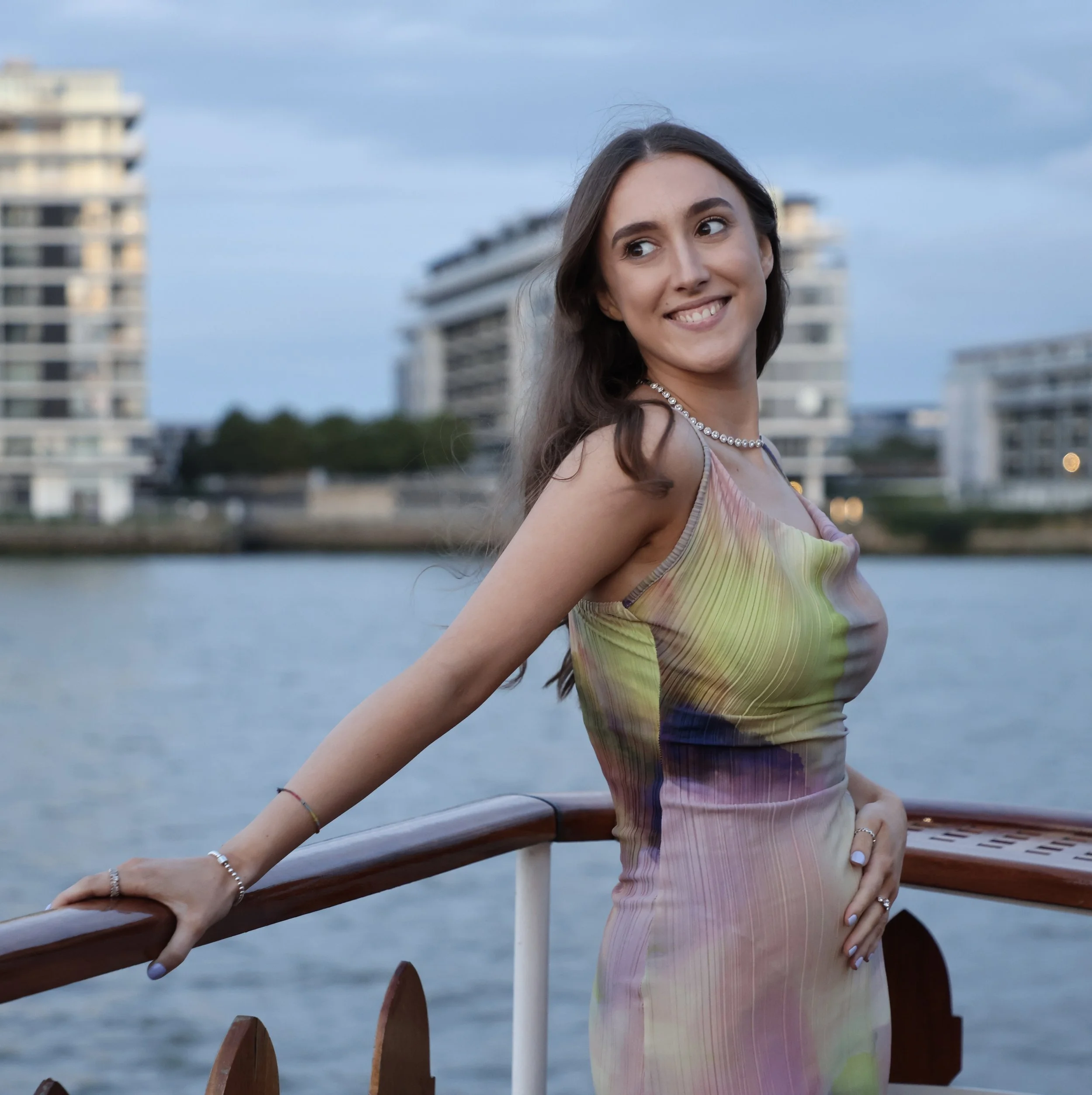 A woman with long brown hair wearing a pink and yellow striped dress and silver jewelry standing on a boat with a wooden railing, smiling and looking to her left, with modern high-rise buildings and a river in the background.