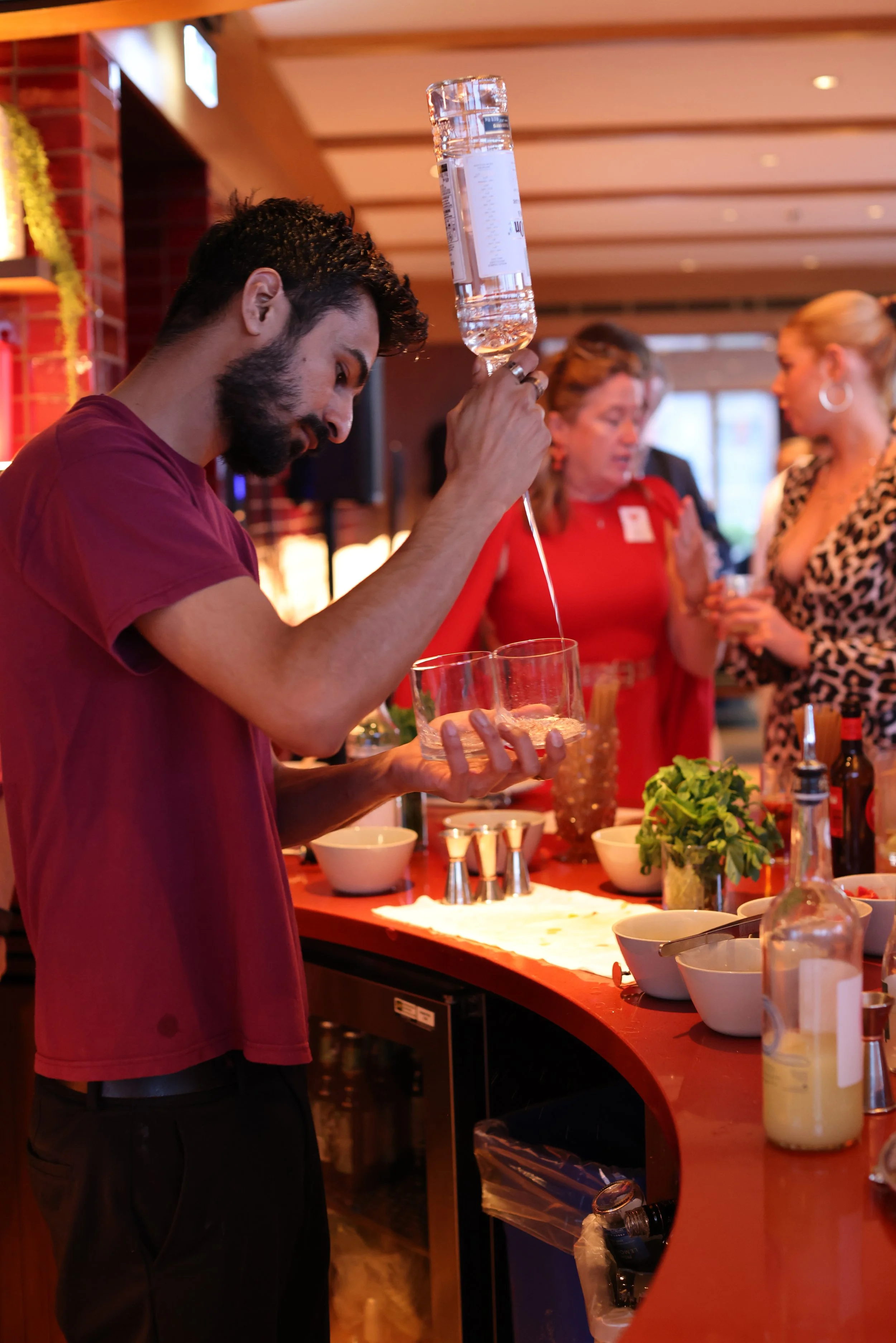 A young man with dark, curly hair, a beard, and wearing a maroon T-shirt is pouring water from a large plastic bottle into a glass. He is standing at a bar or counter with bowls, bottles, and garnishments, with two women in the background dressed in 