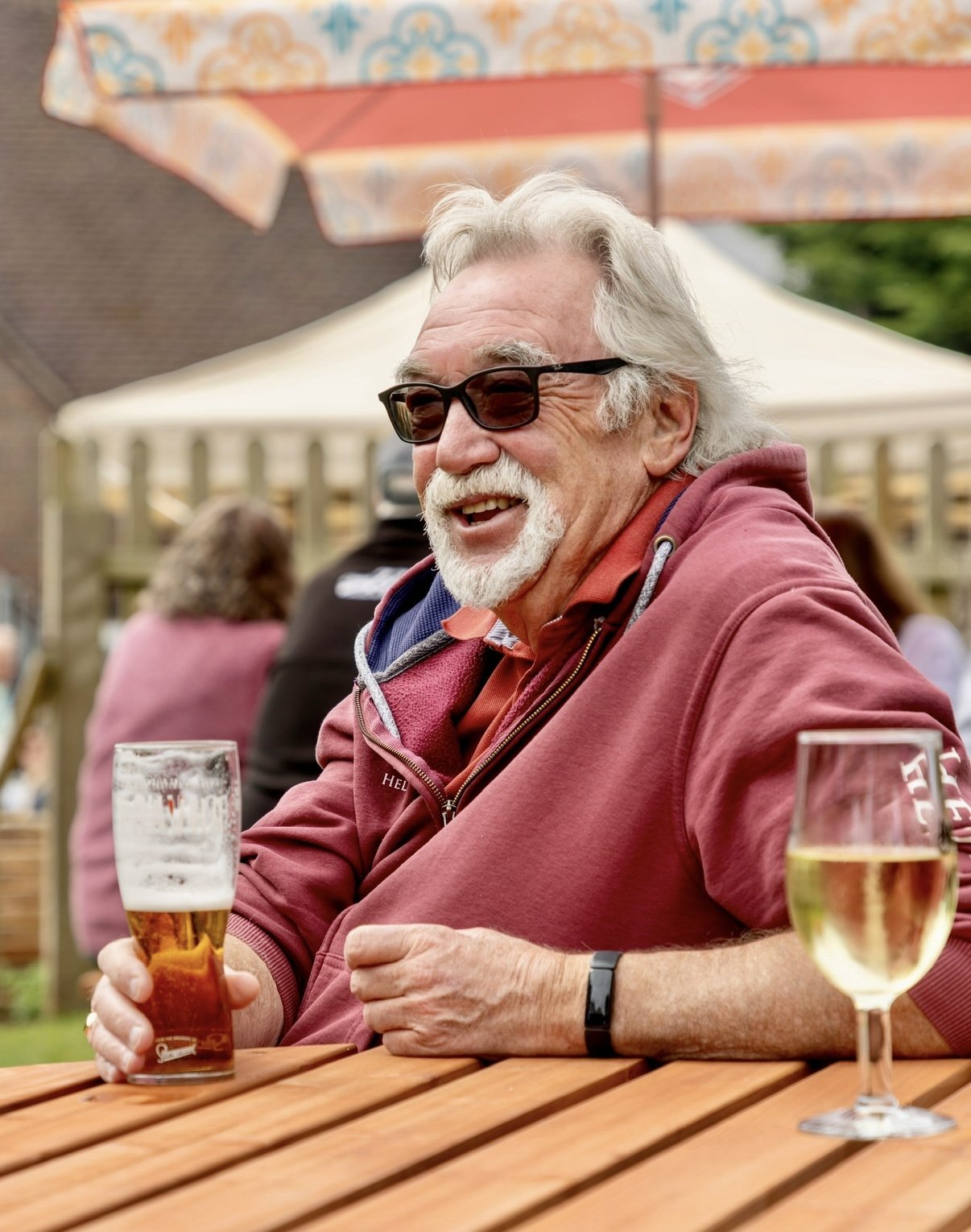 An older man with white hair, beard, and sunglasses smiling while holding a pint of beer at an outdoor gathering. Other people and tables are visible in the background.