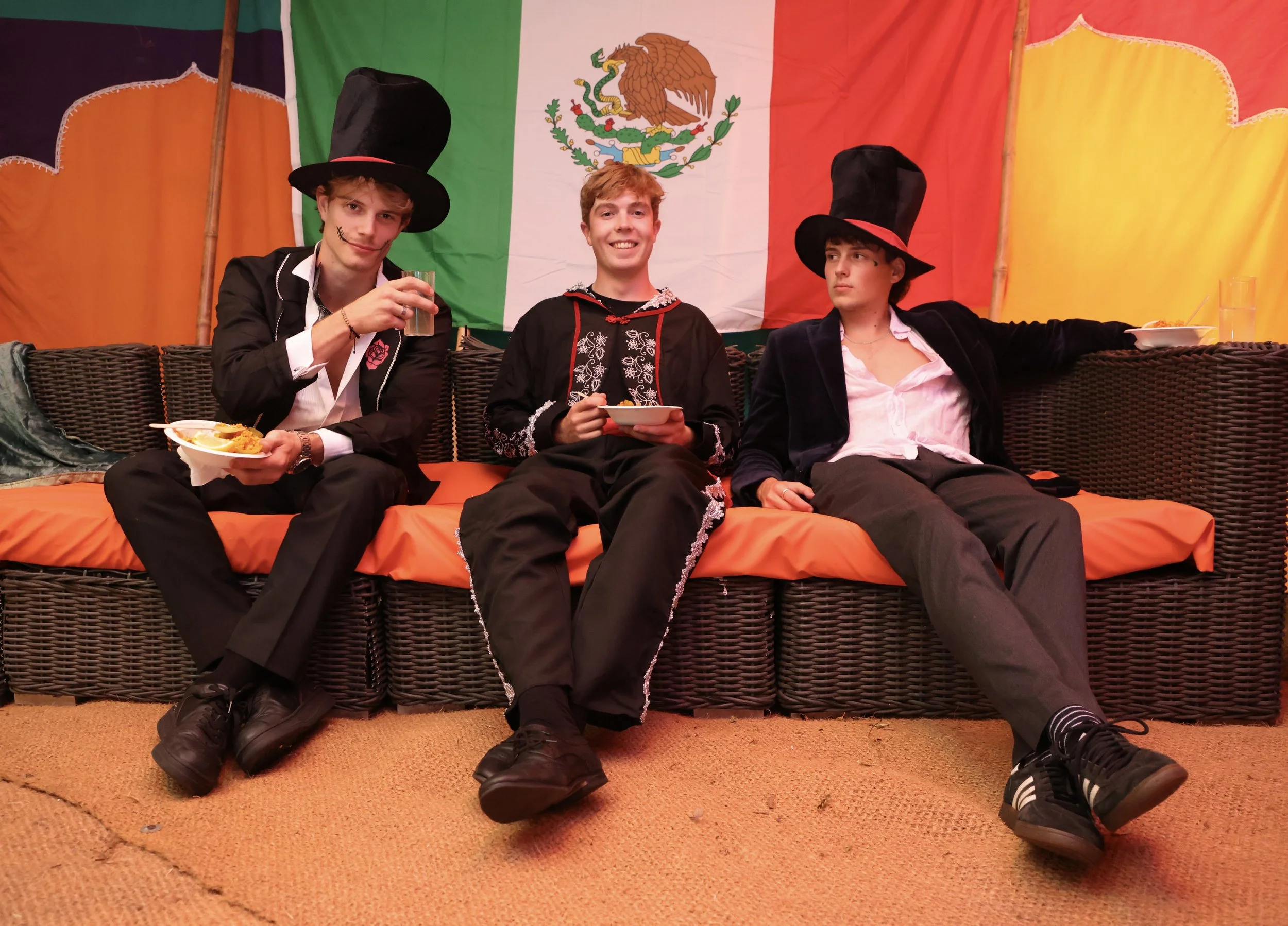 Three young men sitting on a wicker couch in front of a Mexican flag with an eagle and snake emblem, wearing suits and large black hats, holding food and drinks, one with face paint, in a festive setting.