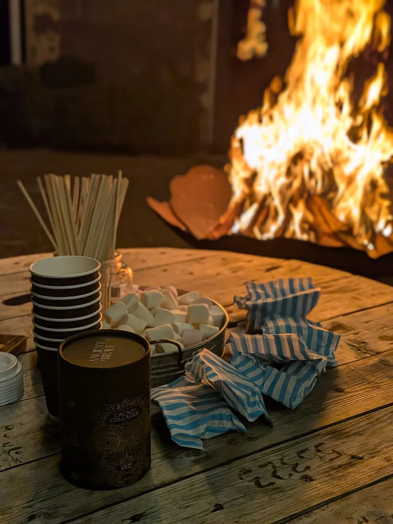 A wooden table with a fire in the background, topped with marshmallows, striped paper wrappers, a tin of cacao and collagen, paper cups, and wooden skewers.